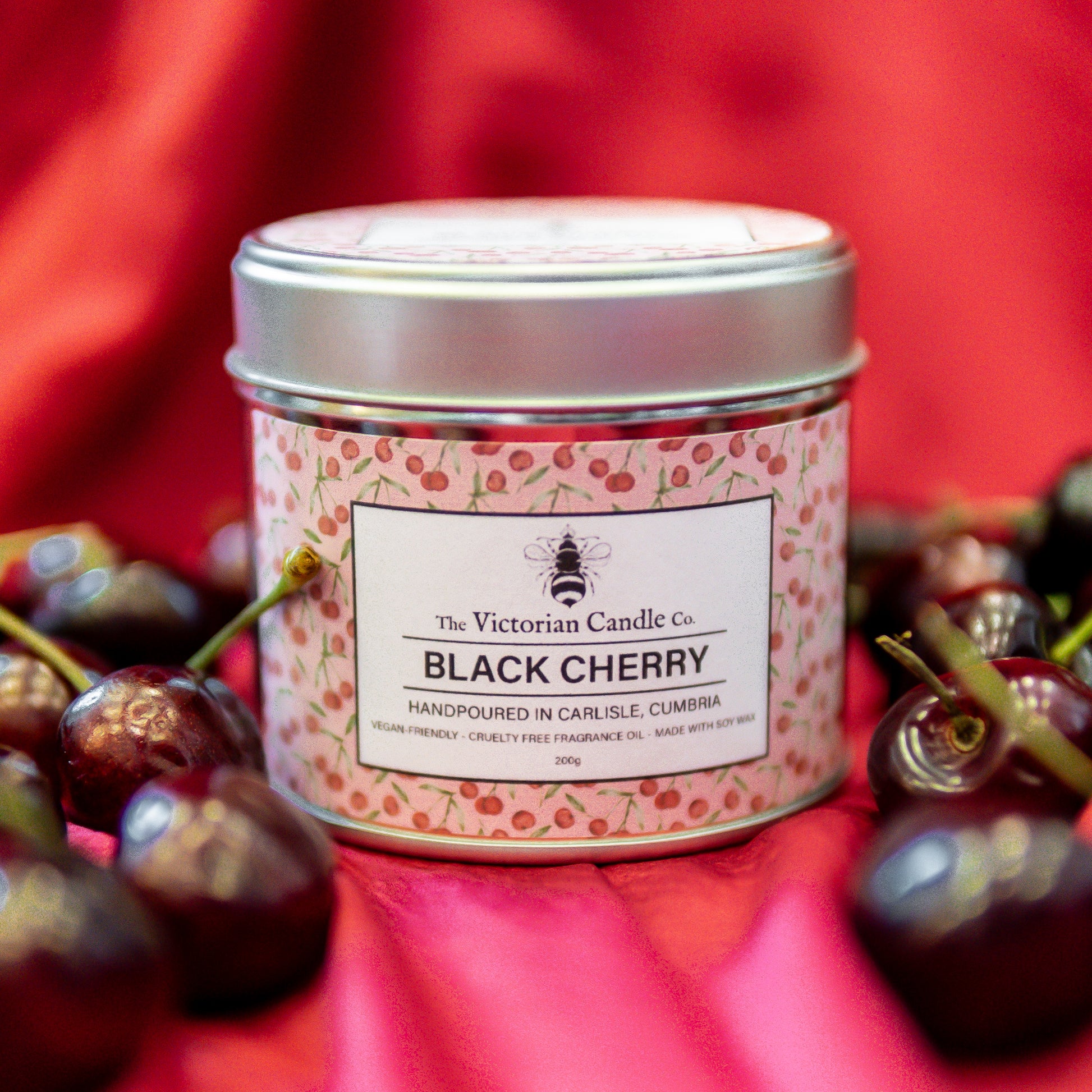 A 200ml tin of The Victorian Candle Co.’s (Large) Black Cherry - Scented Candle sits among fresh cherries on red satin—an ideal Mother’s Day gift. The silver tin’s label shows a cherry pattern and key product details.