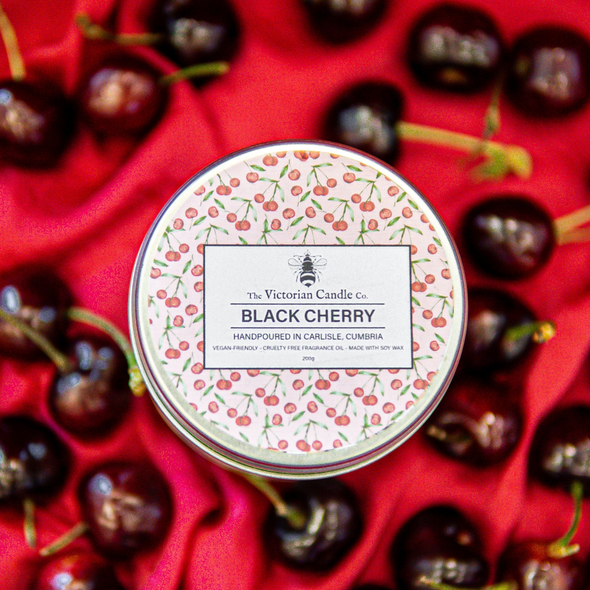 A 200ml tin of The Victorian Candle Co. (Large) Black Cherry Scented Candle rests on red fabric with cherries, featuring a cherry-patterned lid and detailed label—an ideal Mothers Day gift.