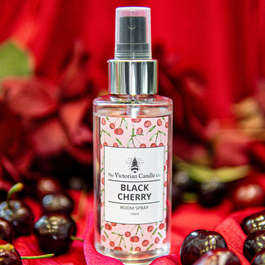 A clear spray bottle labeled Black Cherry - Room Spray from The Victorian Candle Co stands among fresh cherries and red petals, set against a vibrant red background. The label features cherry illustrations, reflecting its bold handmade black cherry fragrance.