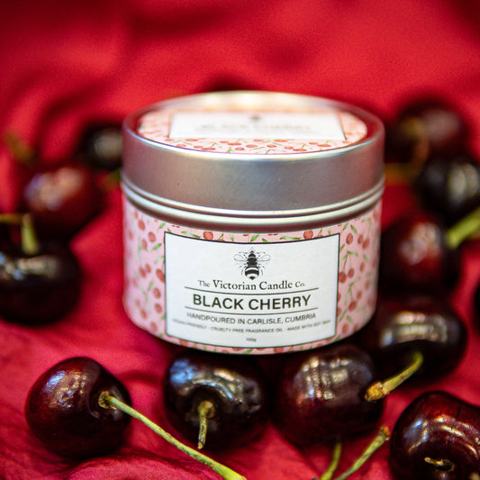 A 100ml tin of The Victorian Candle Co. “Black Cherry” Scented Candle, made with soy vegan-friendly wax, rests on a red cloth beside fresh cherries. Its label is decorated with appealing cherry illustrations.