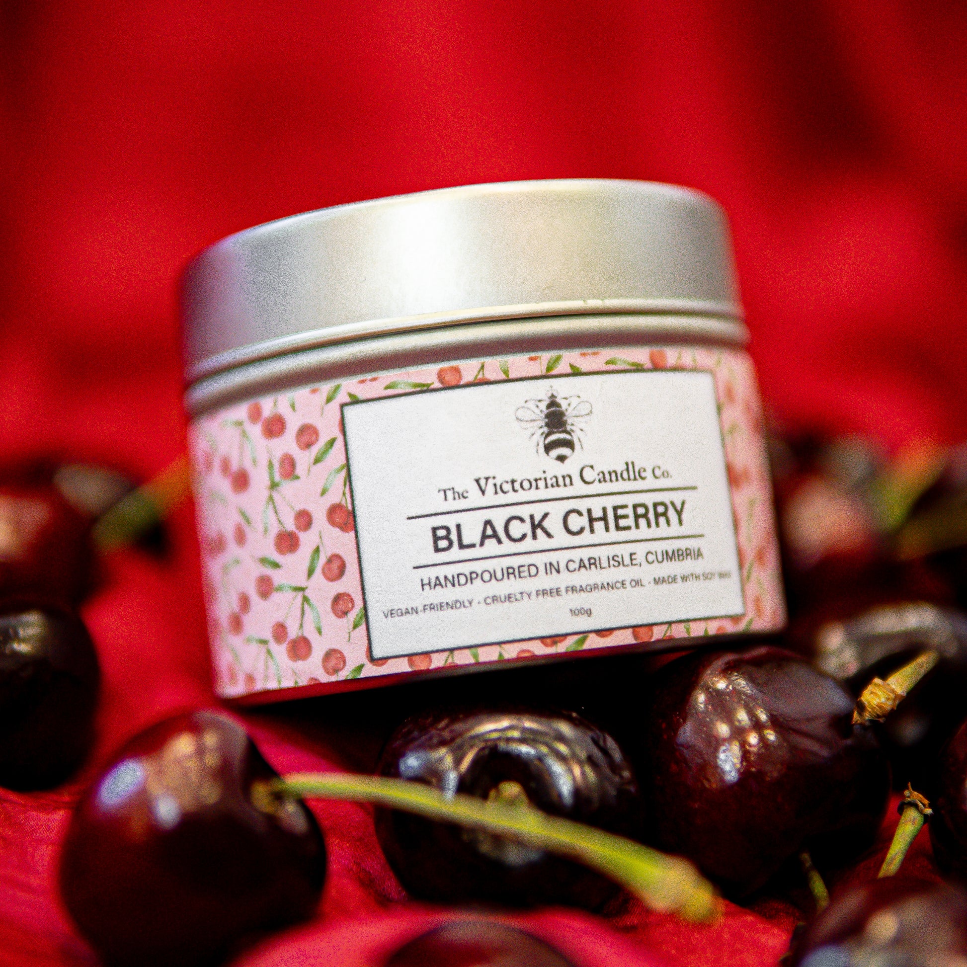A 100ml tin of Black Cherry Scented Candle by The Victorian Candle Co. sits among fresh cherries. Handmade in Carlisle with soy vegan-friendly wax, it features a cherry-patterned label and exudes dark black cherry notes.