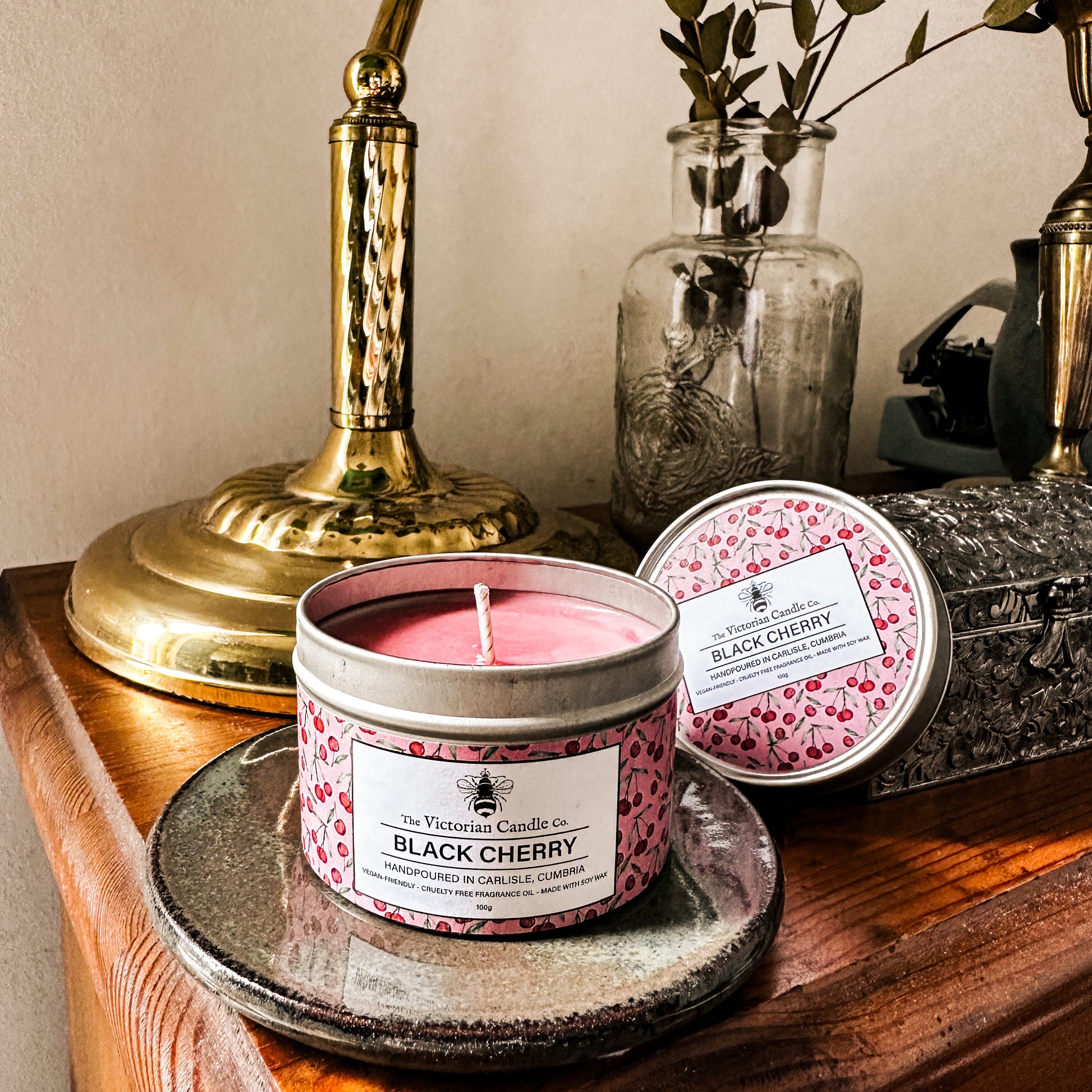 A 100ml Black Cherry Scented Candle tin by The Victorian Candle Co. sits on a dish on a wooden table beside a brass lamp, silver box, and jar with dried leaves. Another matching candle tin appears in the background.