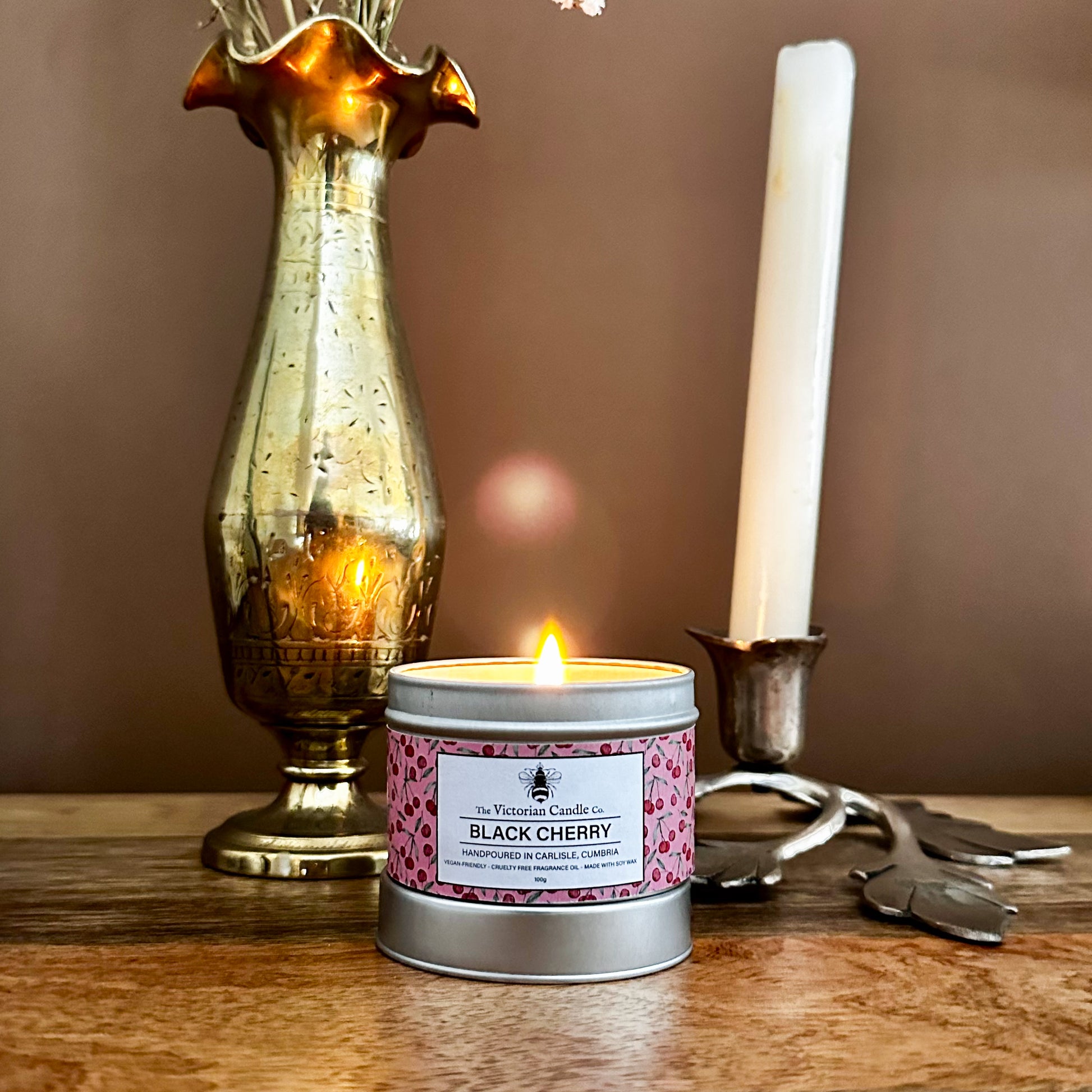 A lit Black Cherry - Scented Candle (100ml Tin) by The Victorian Candle Co. rests on a wooden surface next to a brass vase and an unlit white taper candle in a leaf-shaped metal holder.