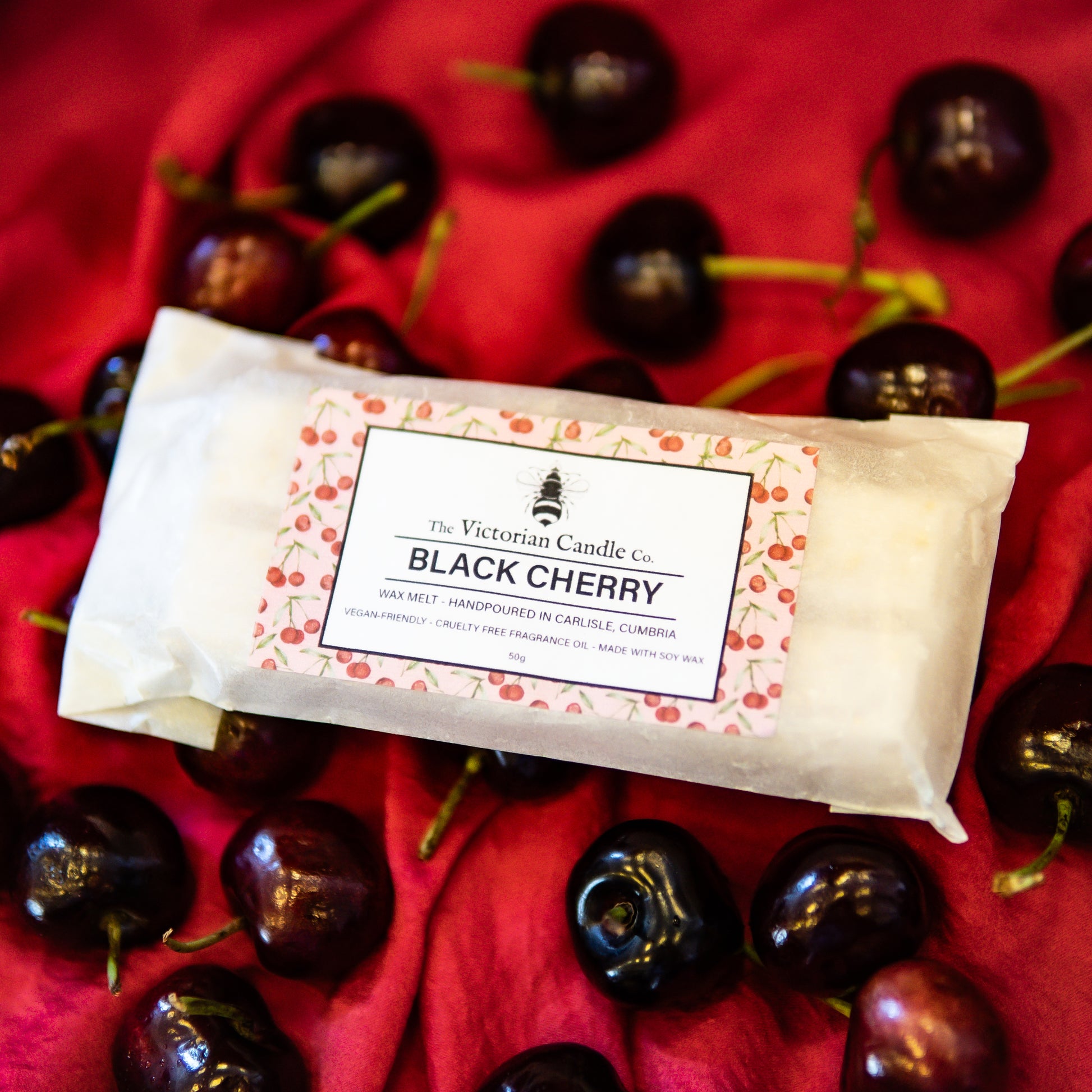 The Victorian Candle Co.s Black Cherry Wax Melt Snap Bar is displayed on red cloth, surrounded by fresh black cherries, evoking a rich and enticing fragrance experience.