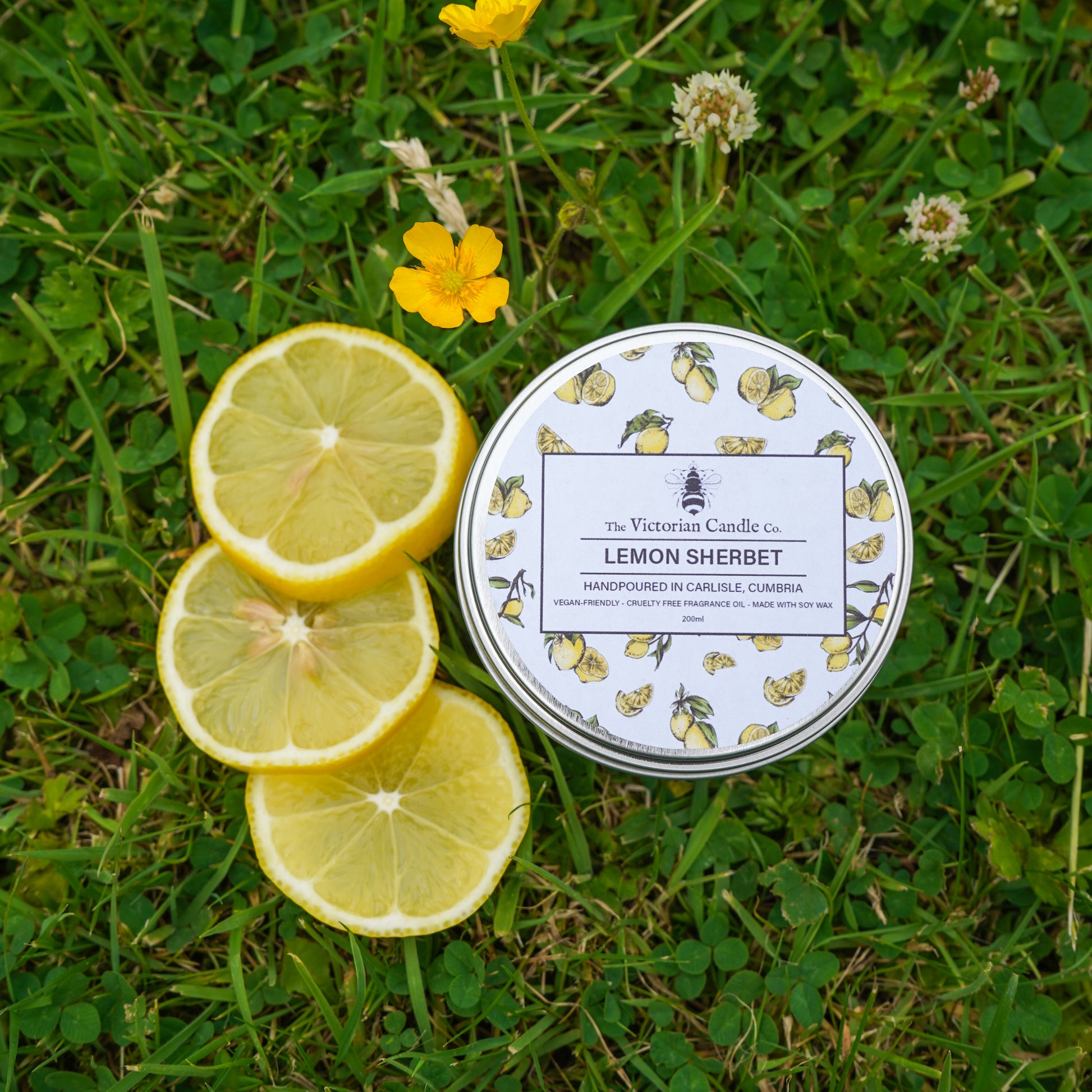 A 200ml tin of The Victorian Candle Co.s (Large) Lemon Sherbet scented candle, featuring a lemon pattern, rests on grass next to three lemon slices and yellow wildflowers.