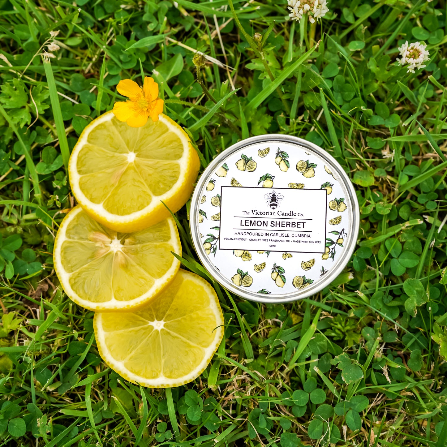 The Victorian Candle Co.s Lemon Sherbet - Scented Candle (100ml tin) with a decorative lemon pattern rests on grass beside three lemon slices and a small yellow flower.