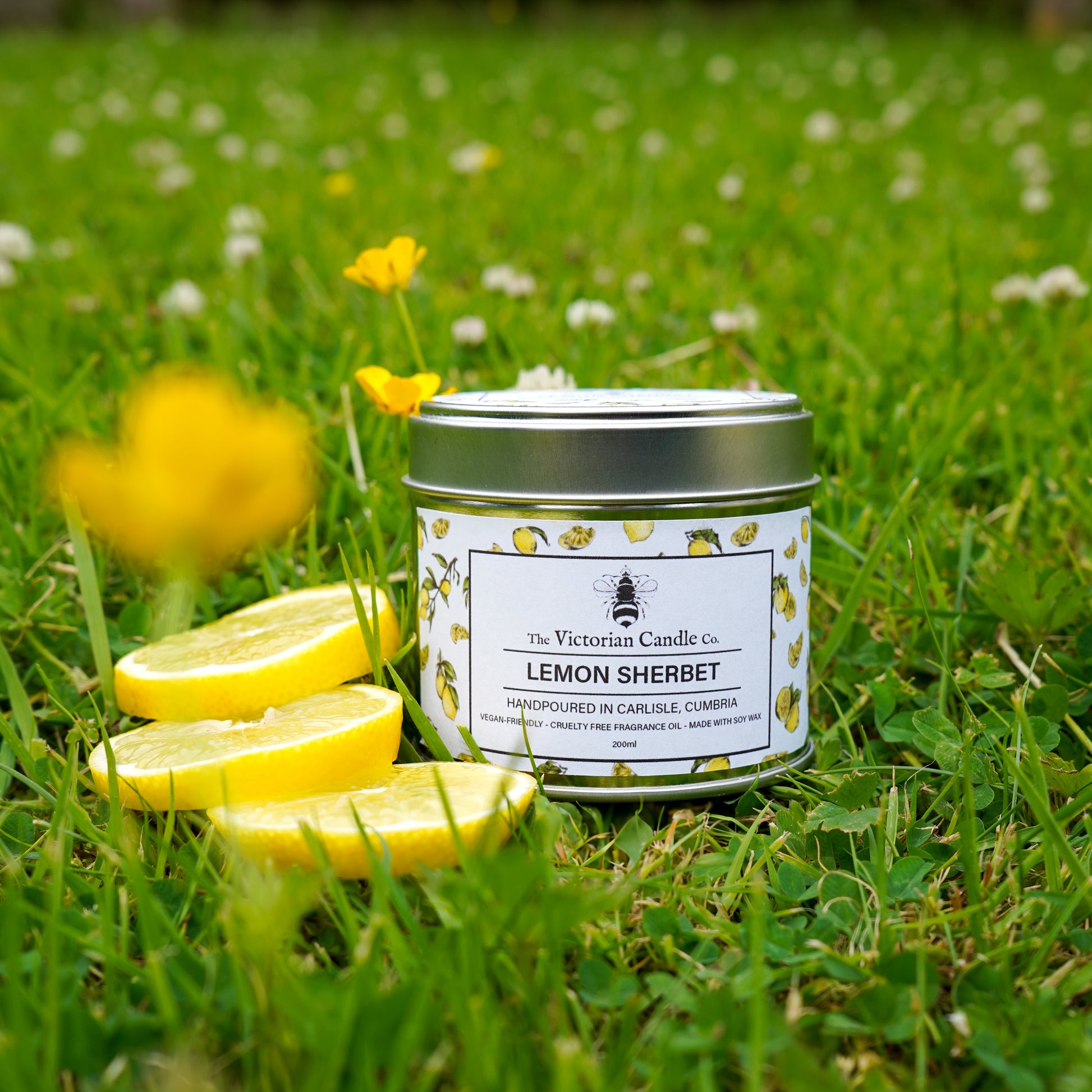 A (Large) Lemon Sherbet - Scented Candle (200ml Tin) by The Victorian Candle Co. rests on green grass with white flowers, alongside three lemon slices and a yellow flower in the foreground.