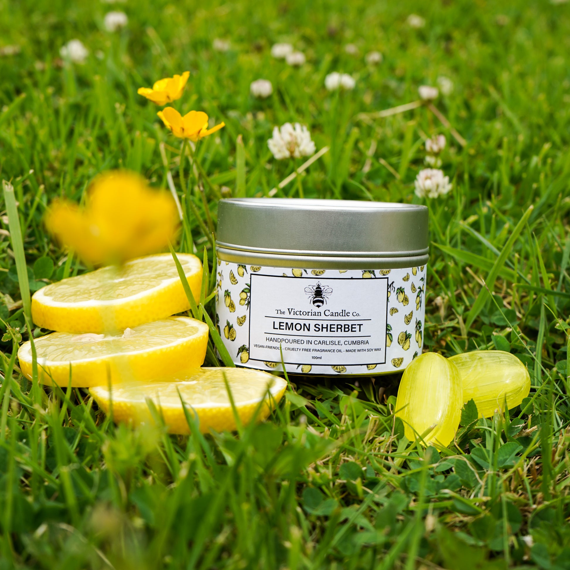 A 100ml tin of The Victorian Candle Co. Lemon Sherbet Scented Candle rests on grass, surrounded by yellow wildflowers, lemon slices, and two yellow wax melts.