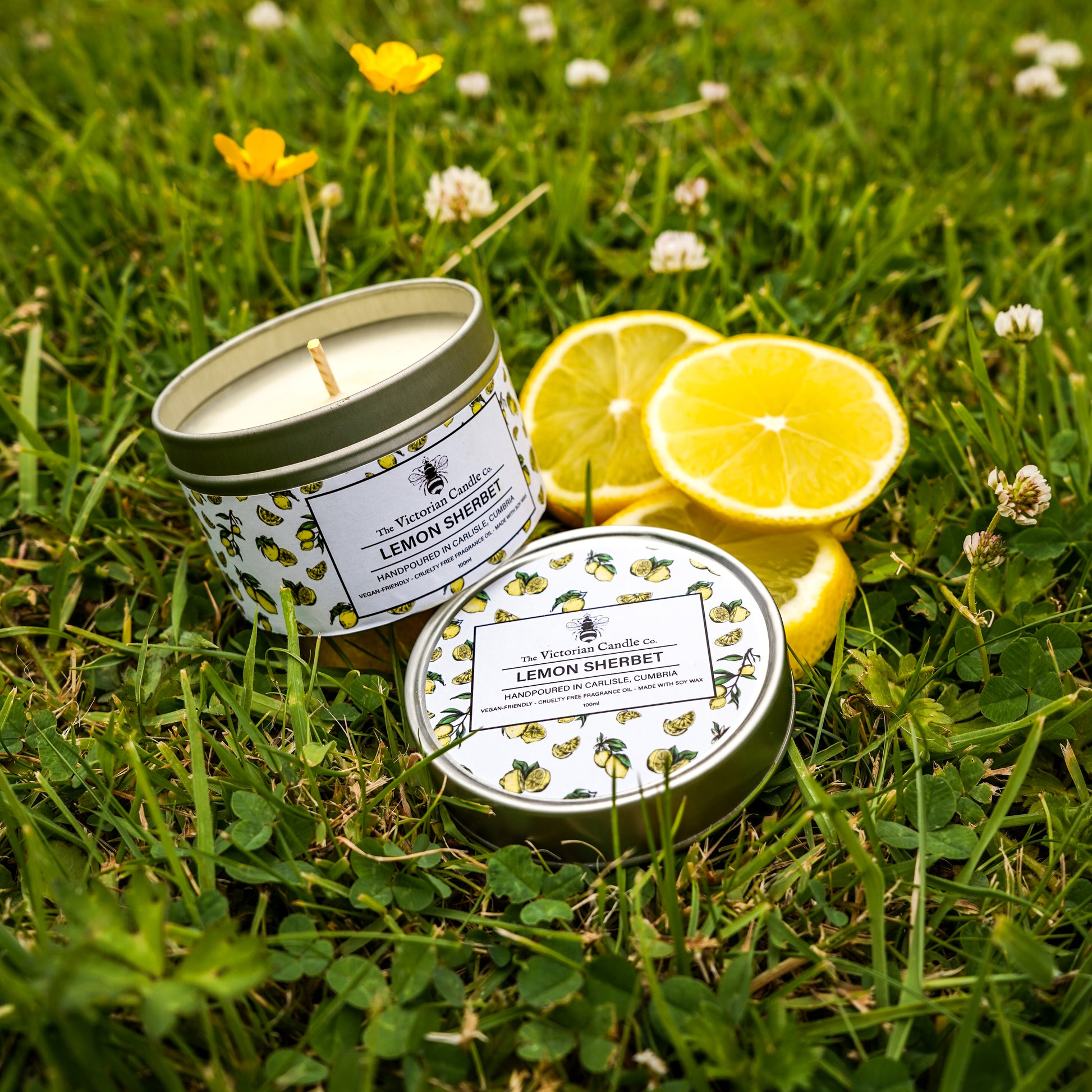 The Victorian Candle Co.s Lemon Sherbet Scented Candle (100ml tin) with a decorative lemon pattern rests open on grass, its lid nearby, alongside fresh lemon slices and small yellow flowers.
