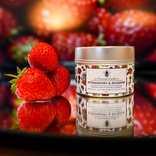 A 100ml tin of The Victorian Candle Co. Strawberry & Rhubarb Scented Candle sits on a reflective surface beside three fresh strawberries, with more strawberries blurred in the background, showcasing this delightful soy wax candle.