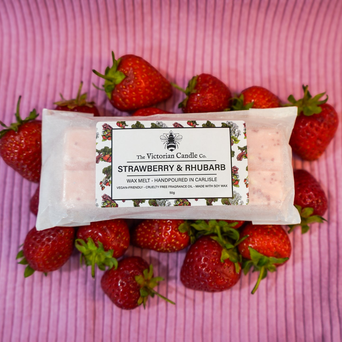 The Victorian Candle Co.s Strawberry & Rhubarb Wax Melt Snap Bar, handmade in Carlisle with vegan-friendly soy wax, sits on a pink striped surface with fresh strawberries, offering a cruelty-free, long-lasting fragrance.