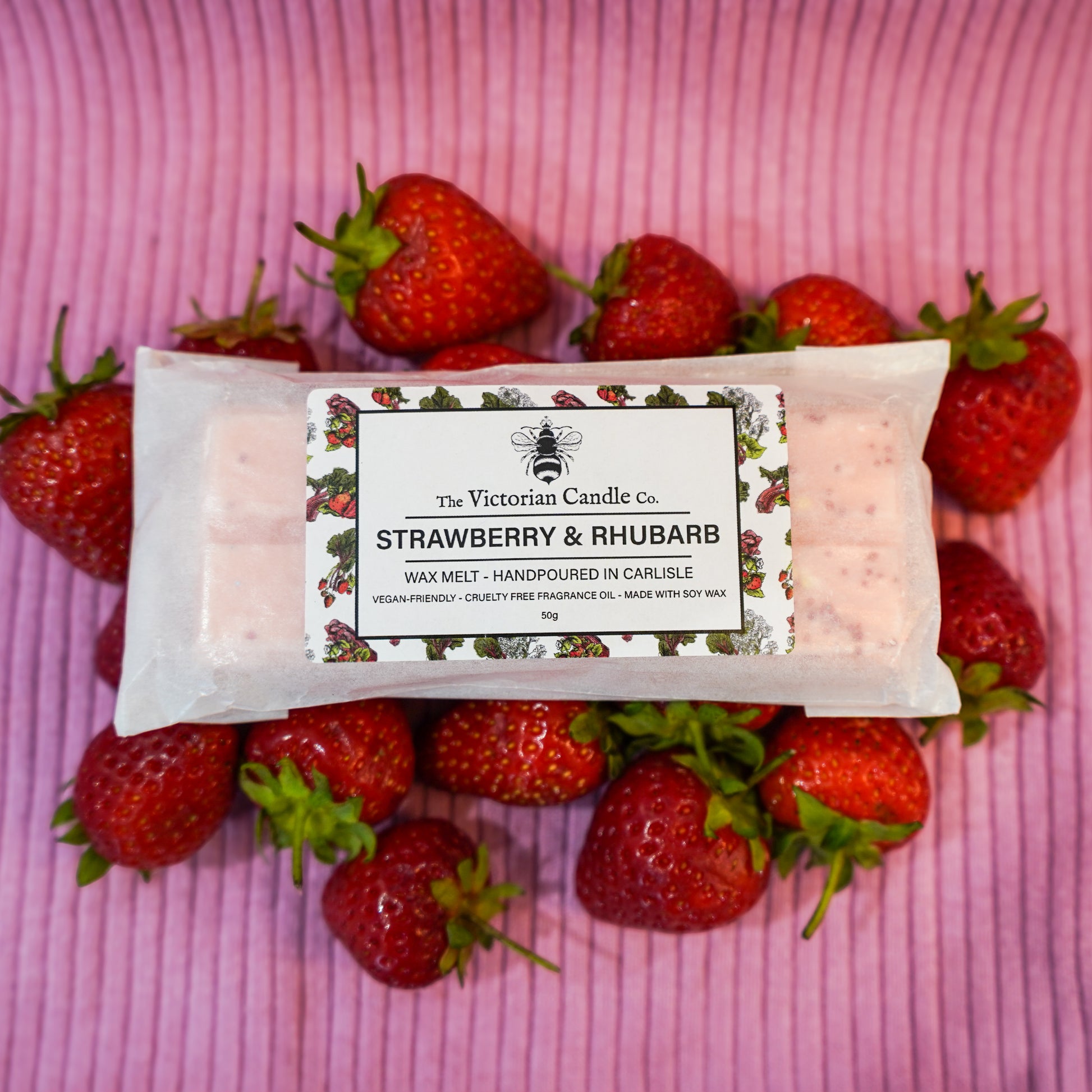 The Victorian Candle Co.s Strawberry & Rhubarb Wax Melt Snap Bar, handmade in Carlisle with vegan-friendly soy wax, sits on a pink striped surface with fresh strawberries, offering a cruelty-free, long-lasting fragrance.