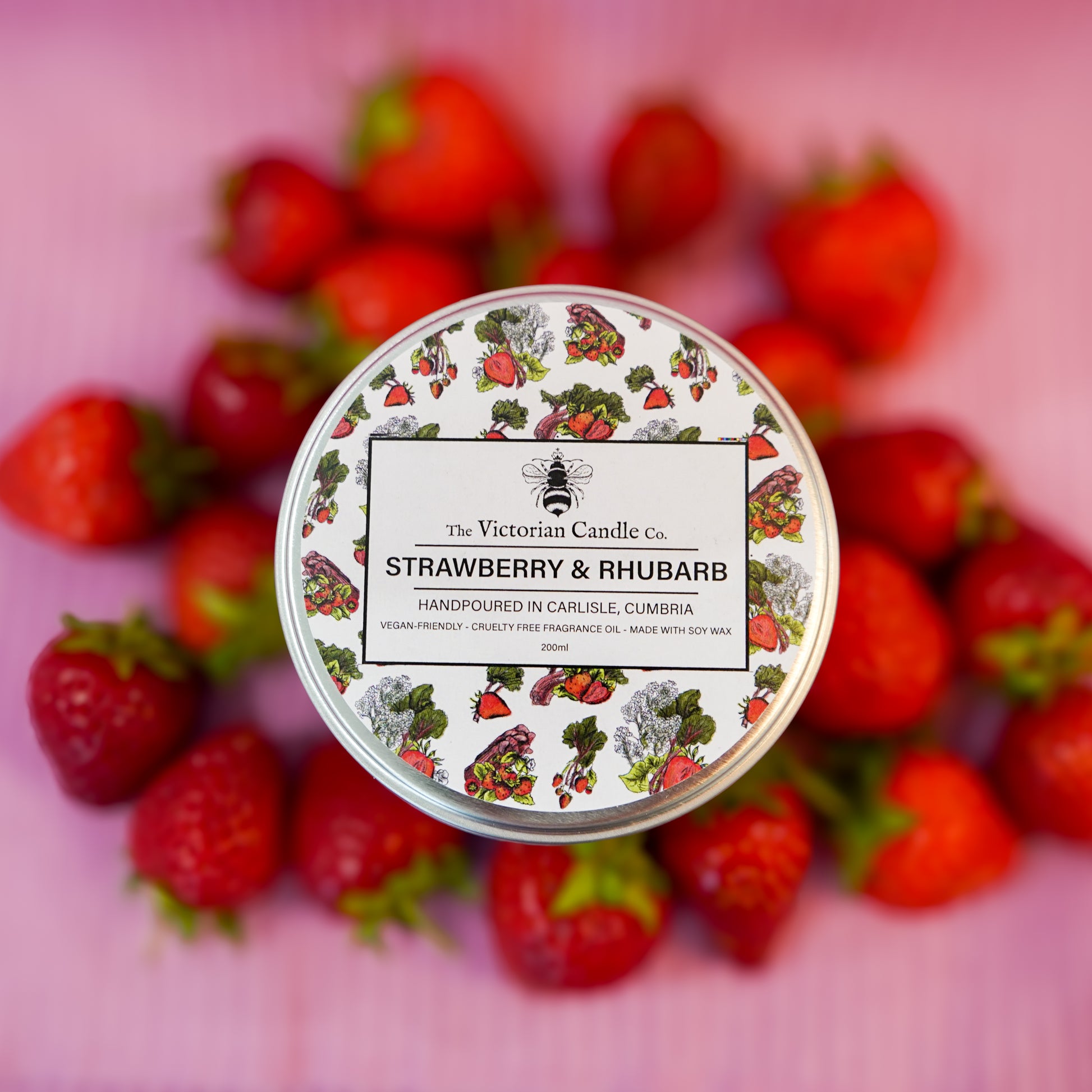 The Victorian Candle Co. (Large) Strawberry & Rhubarb Scented Candle—200ml, a vegan soy wax candle with strawberry and rhubarb illustrations on the tin, rests on a pink surface surrounded by fresh strawberries.