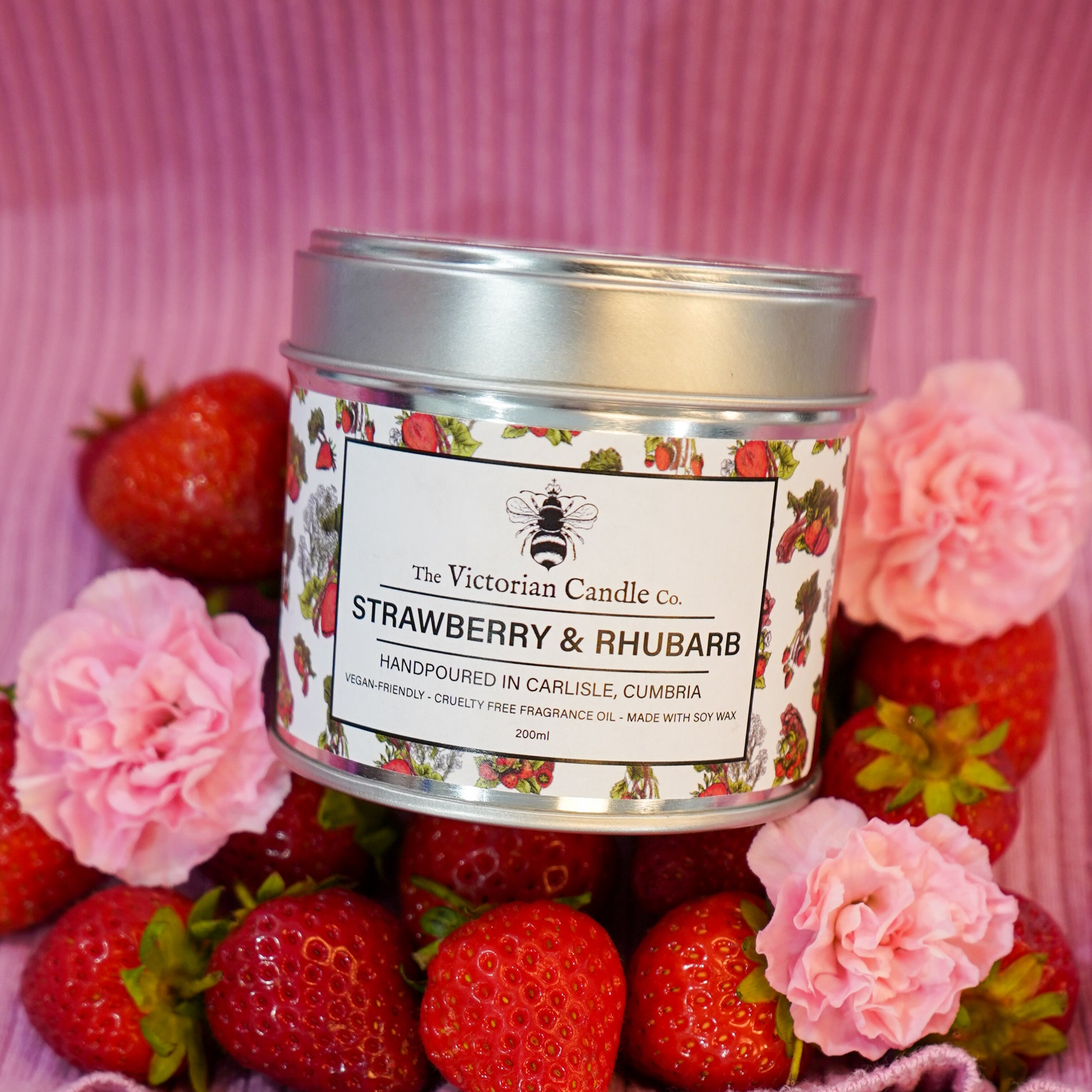 A 200ml tin of The Victorian Candle Co. (Large) Strawberry & Rhubarb Scented Candle sits among fresh strawberries and pink flowers on a pink striped background, with a vegan-friendly bee and floral label design.