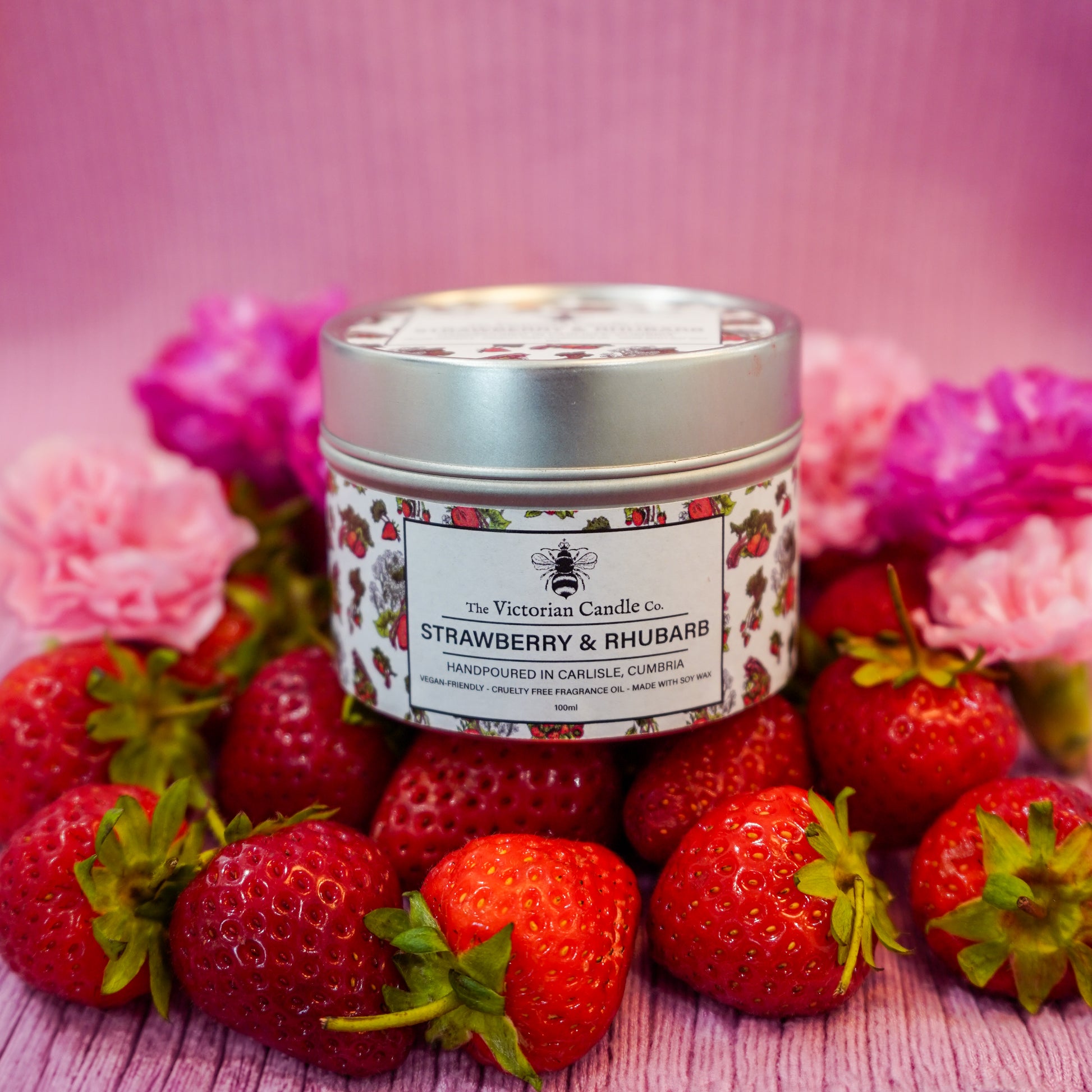 The Victorian Candle Co. Strawberry & Rhubarb - Scented Candle (100ml tin) is displayed among fresh strawberries and pink flowers on a pink striped background—an ideal way to showcase this delightful soy wax candle.