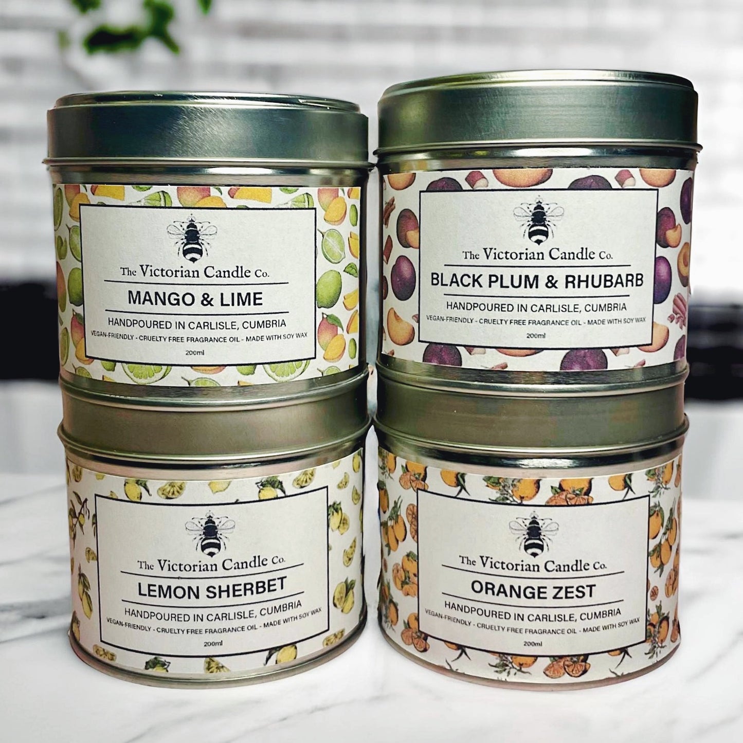 Four tins from The Victorian Candle Co.'s Fruity Large Candle Collection Box - 200ml Tins are stacked on a white surface against a tiled background. This collection features fruity scents such as Mango & Lime, Black Plum & Rhubarb, Lemon Sherbet, and Orange Zest, all crafted from soy wax for a cleaner burn.