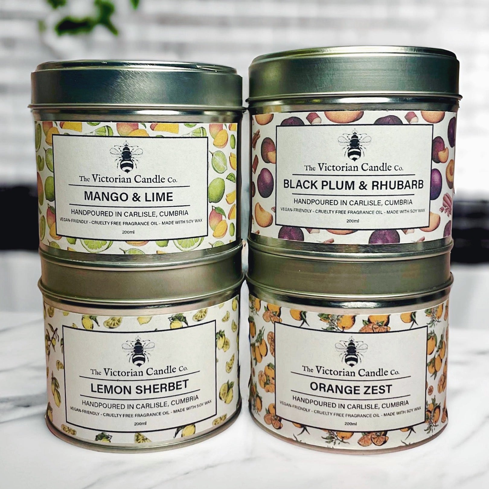 Four tins from The Victorian Candle Co.'s Fruity Large Candle Collection Box - 200ml Tins are stacked on a white surface against a tiled background. This collection features fruity scents such as Mango & Lime, Black Plum & Rhubarb, Lemon Sherbet, and Orange Zest, all crafted from soy wax for a cleaner burn.