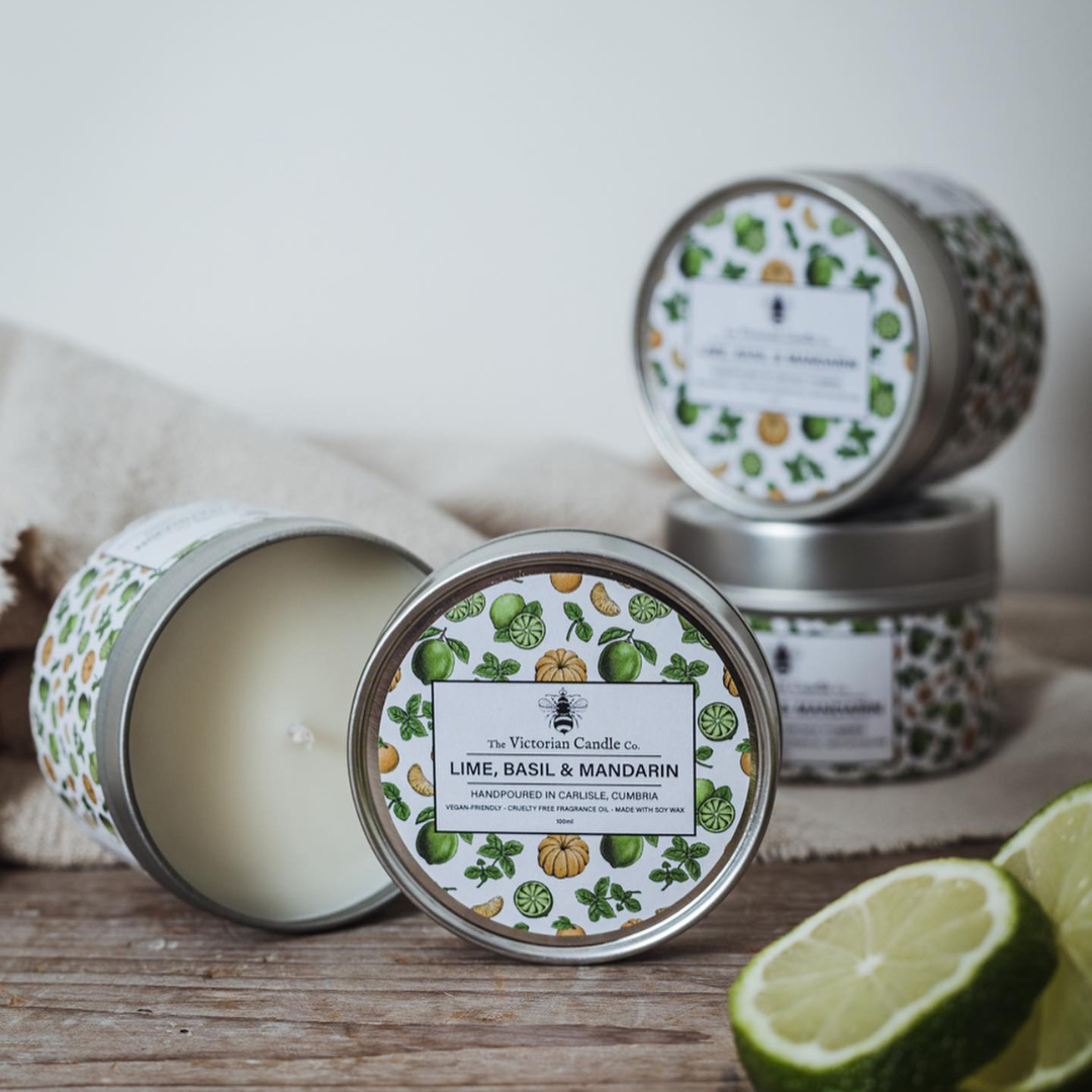 A display of The Victorian Candle Co.'s Lime, Basil & Mandarin Scented Candles in 100ml tins showcases their decorative allure. One candle is open, revealing the sleek white wax within. Nearby, sliced limes and a beige cloth enhance the fresh and inviting aroma of these soy vegan-friendly products.