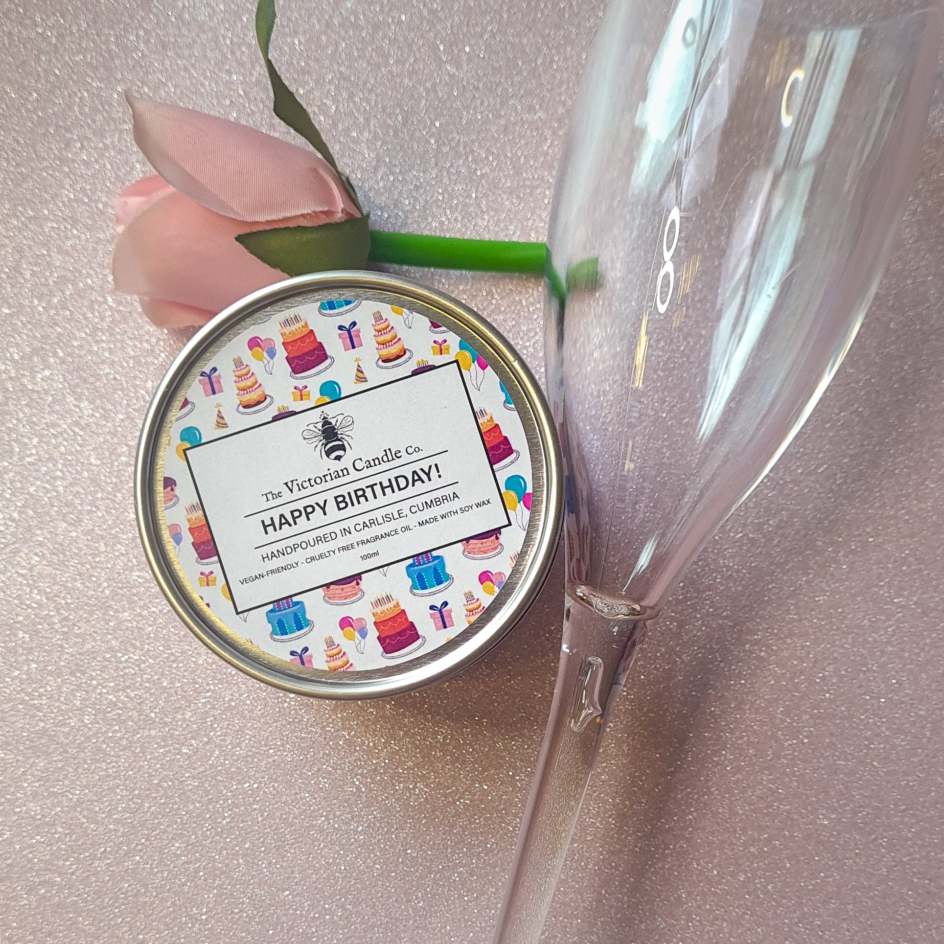 A soy wax candle labeled as "Happy Birthday - Scented Candle - 100ml Tin" by The Victorian Candle Co., featuring a festive design, rests on a glittery surface. Accompanied by a single pink rose and a clear champagne flute, it adds to the celebratory arrangement with its delightful aroma.