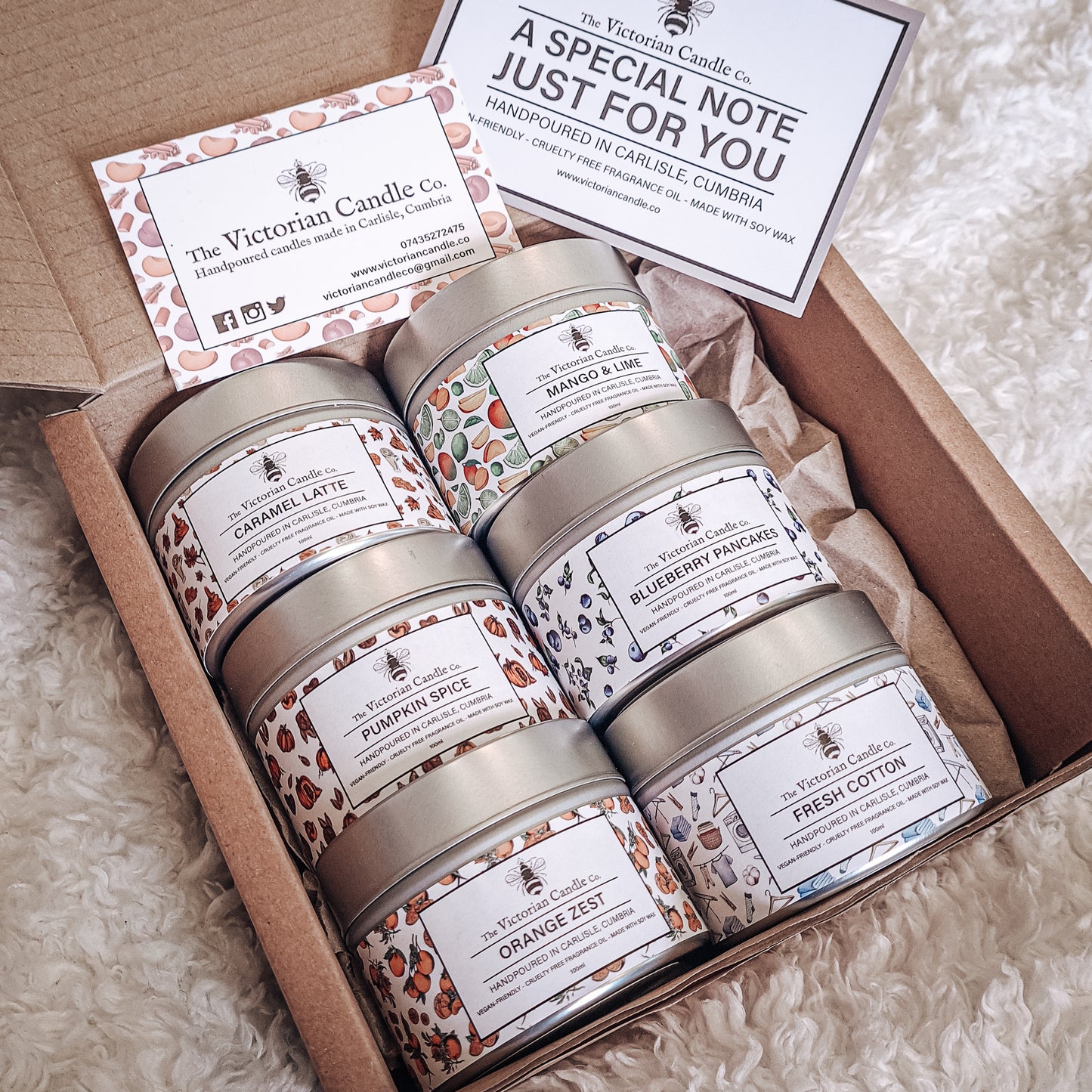 The 6 Small Candle Gift Box 🎁 from The Victorian Candle Co. features six scented candles in tins, each labeled with different scents such as Caramel Latte and Fresh Cotton. Accompanied by a card with a personalized message from The Victorian Candle Co., all items are elegantly arranged on a textured white surface—ideal for any occasion as a beautifully crafted handmade gift.