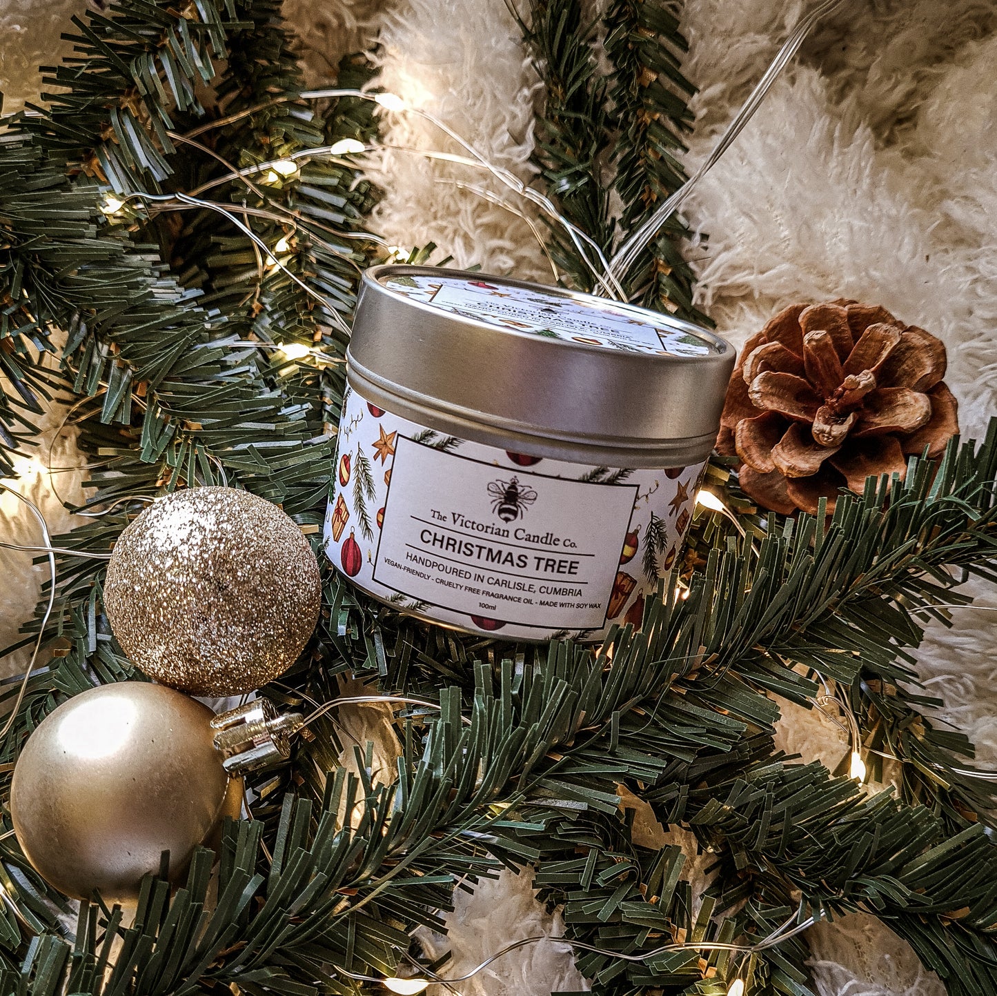 A cheerful arrangement showcasing The Victorian Candle Co.'s Christmas Tree - Scented Candle in a 100ml Tin 🎄, placed amidst lush evergreen branches, a pine cone, radiant warm white string lights, and two sparkling gold ornaments on a plush, white surface.