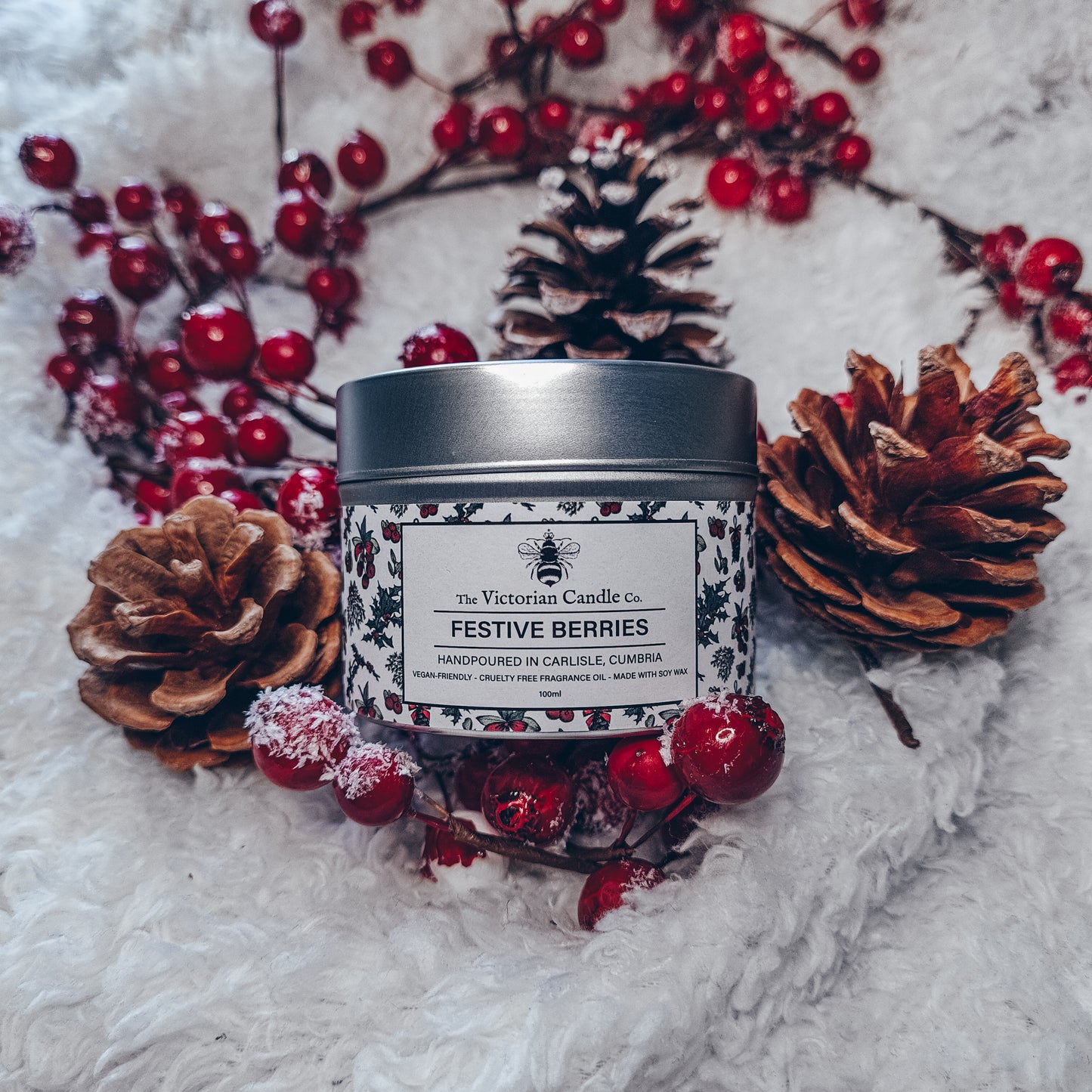 A Festive Berries - Scented Candle - 100ml Tin 🎅🏻 from The Victorian Candle Co. rests on a fluffy white surface, adorned with pine cones and red berries. Crafted from soy vegan-friendly wax, its cranberry scent envelops the air, evoking a warm holiday atmosphere.