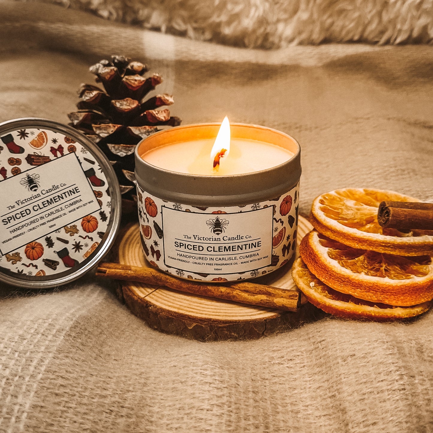 A Spiced Clementine - Scented Candle - 100ml Tin by The Victorian Candle Co. rests on a cozy blanket, accompanied by dried orange slices, cinnamon sticks, and a pine cone. The candle's decorative lid, adorned with autumn leaves and pumpkins, is propped against it.