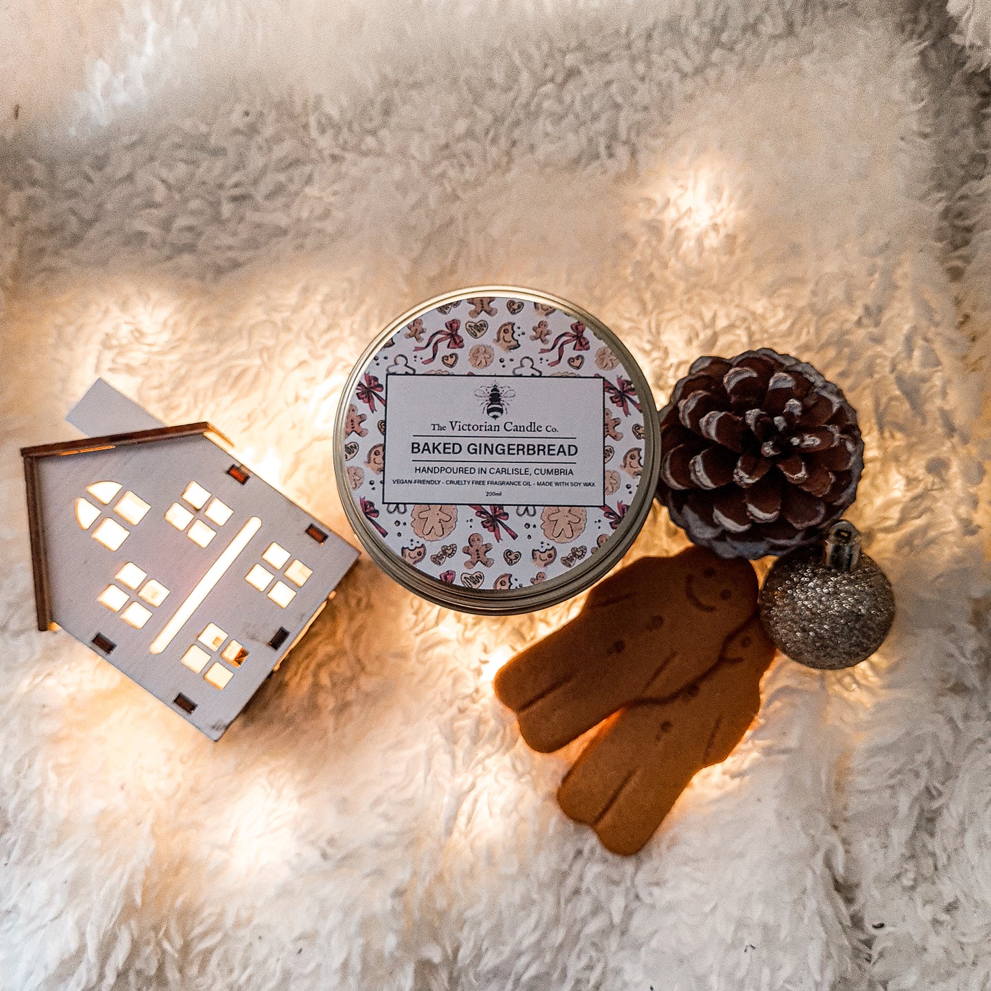 A cozy scene featuring a lit (Limited Edition) Large Baked Gingerbread Scented Candle - 200ml Gold Tin by The Victorian Candle Co., accompanied by a small decorative house with lights, a pinecone, a gingerbread cookie, and a glittery ornament all arranged on a fluffy white blanket.