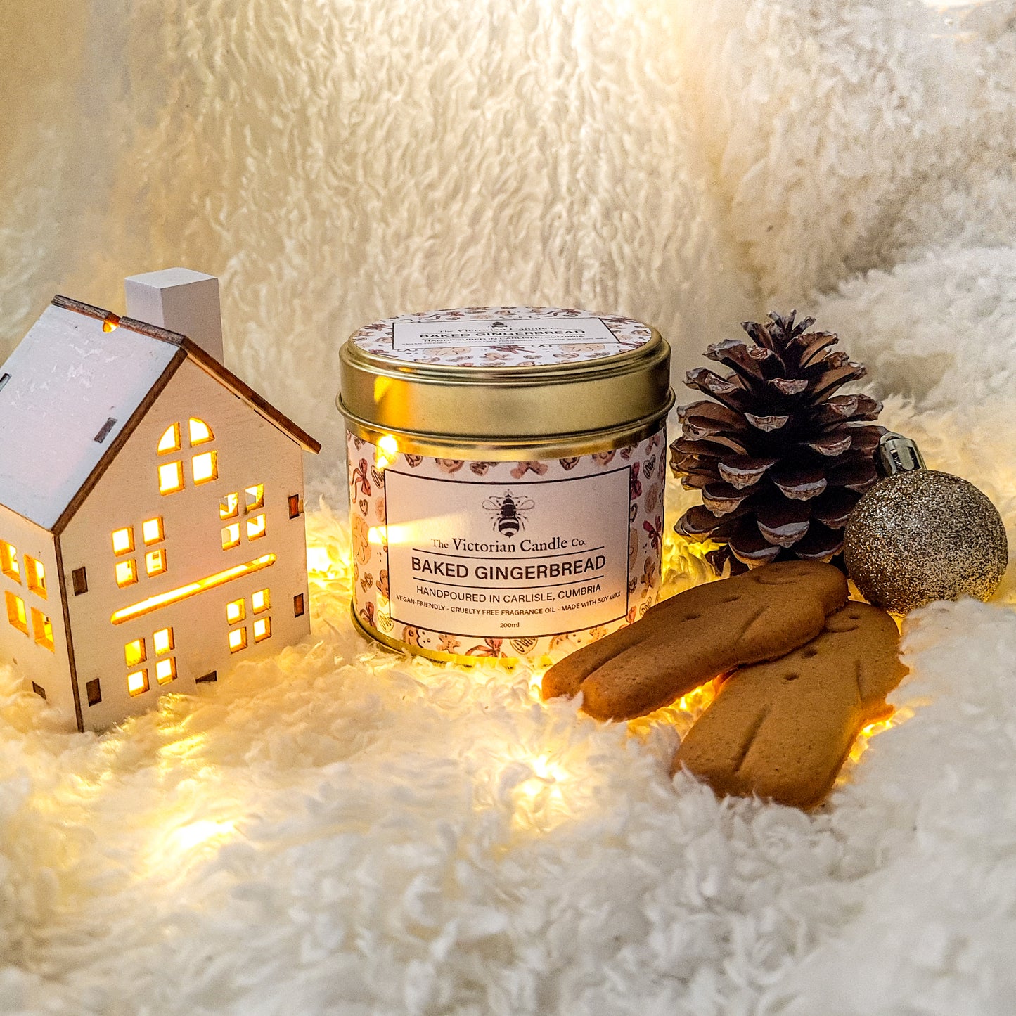 Imagine a cozy scene featuring a lit Limited Edition Large Baked Gingerbread Scented Candle in a 200ml Gold Tin by The Victorian Candle Co., surrounded by a decorative white ceramic house with glowing windows, a pine cone, gingerbread cookies, and a glittery ornamentāall set elegantly on a fluffy white surface.