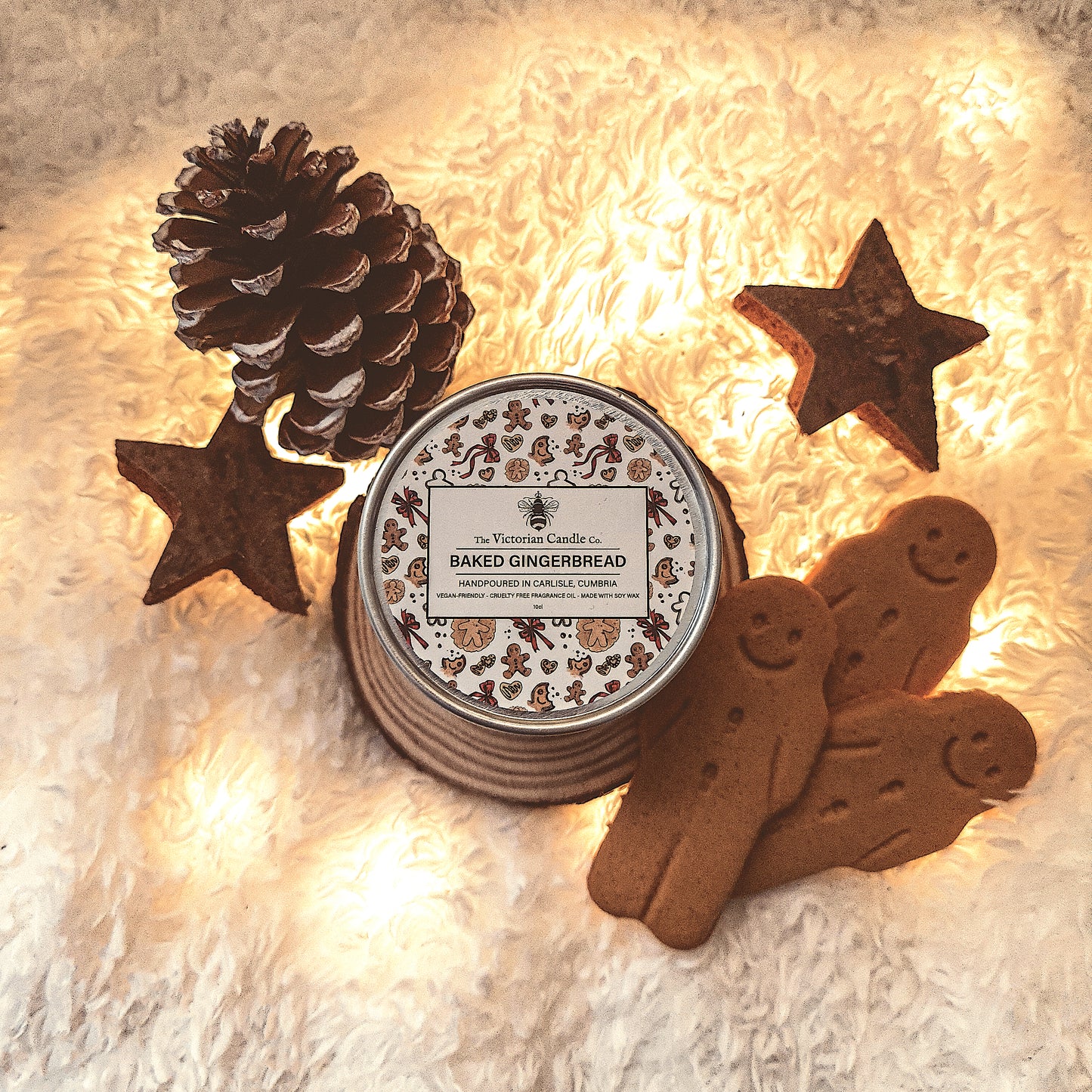 A cozy arrangement features a Baked Gingerbread - Scented Candle - 100ml Tin 🍪 from The Victorian Candle Co., casting a warm glow over two gingerbread cookies, star-shaped ornaments, and a pine cone on a fluffy white surface. This candle enriches the festive, wintry atmosphere with its inviting aroma.