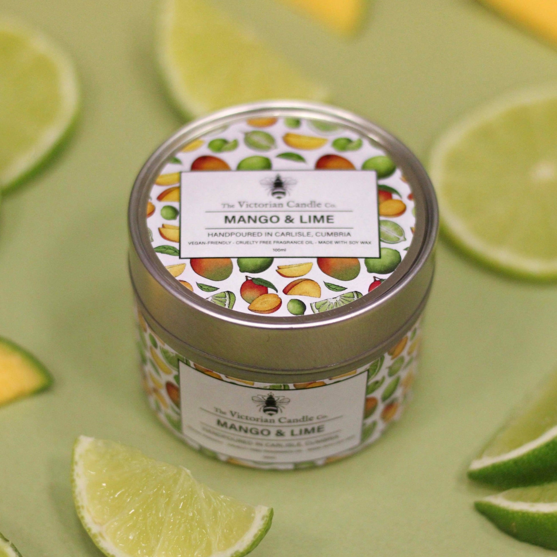 A decorative tin labeled Mango & Lime - Scented Candle - 100ml Tin from The Victorian Candle Co. rests on a green surface, showcasing vegan-friendly illustrations of mangos and limes, beautifully accented by slices of fresh lime scattered around it.