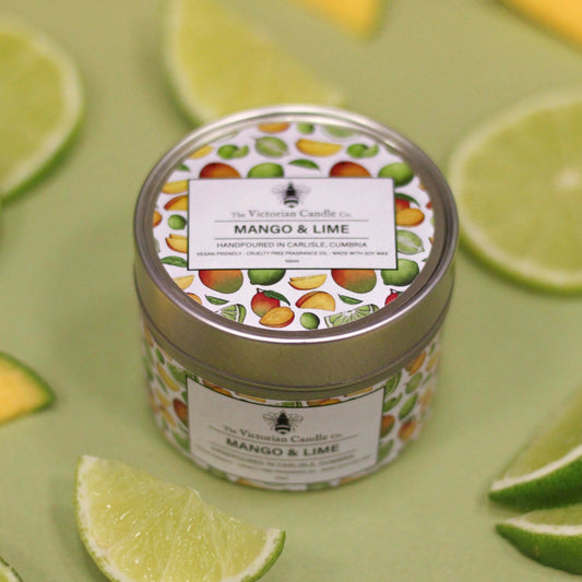 A decorative tin labeled Mango & Lime - Scented Candle - 100ml Tin from The Victorian Candle Co. rests on a green surface, showcasing vegan-friendly illustrations of mangos and limes, beautifully accented by slices of fresh lime scattered around it.
