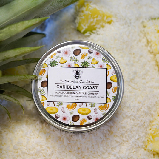 A round 100ml tin of the Caribbean Coast Scented Candle by The Victorian Candle Co. sits on a layer of shredded coconut, releasing its tropical scent. The label features vibrant illustrations of fruit slices and leaves, with a touch of pineapple in the top left corner. Crafted from soy vegan-friendly wax for an eco-conscious experience.