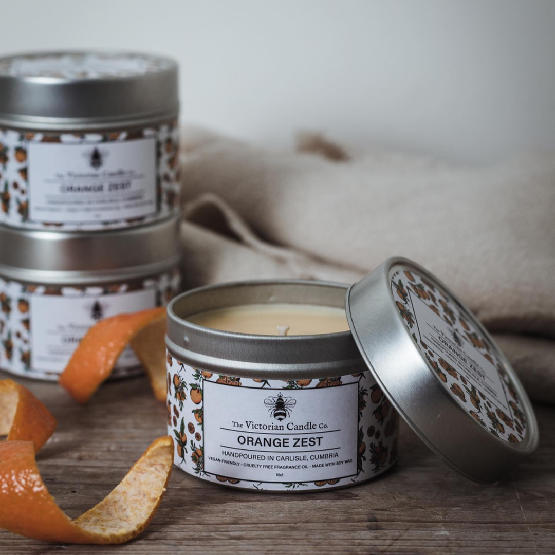 An open 100ml tin of Orange Zest Scented Candle by The Victorian Candle Co. reveals the candle inside, crafted from soy vegan-friendly wax. The recyclable tin, adorned with a floral design, sits near several orange peels. In the background, more tins are stacked on a soft fabric surface.
