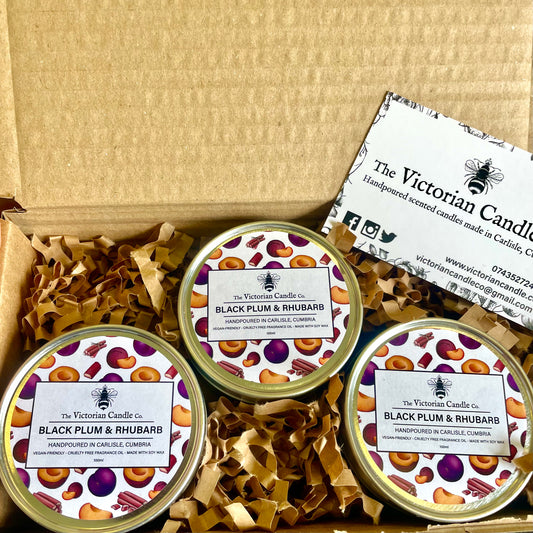 A Trio Box featuring three 100ml tins of Black Plum & Rhubarb scented candles, each adorned with decorative fruit labels, is presented in an open box lined with brown crinkle paper. These soy wax candles are accompanied by a business card from The Victorian Candle Co.