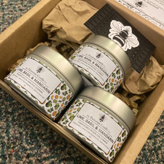The Trio Box from The Victorian Candle Co., featuring three 100ml tins of Lime, Basil & Mandarin scented candles, is elegantly packaged with tissue paper. Crafted from soy wax, the set includes a card adorned with a bee illustration and is placed on a speckled carpet for display.