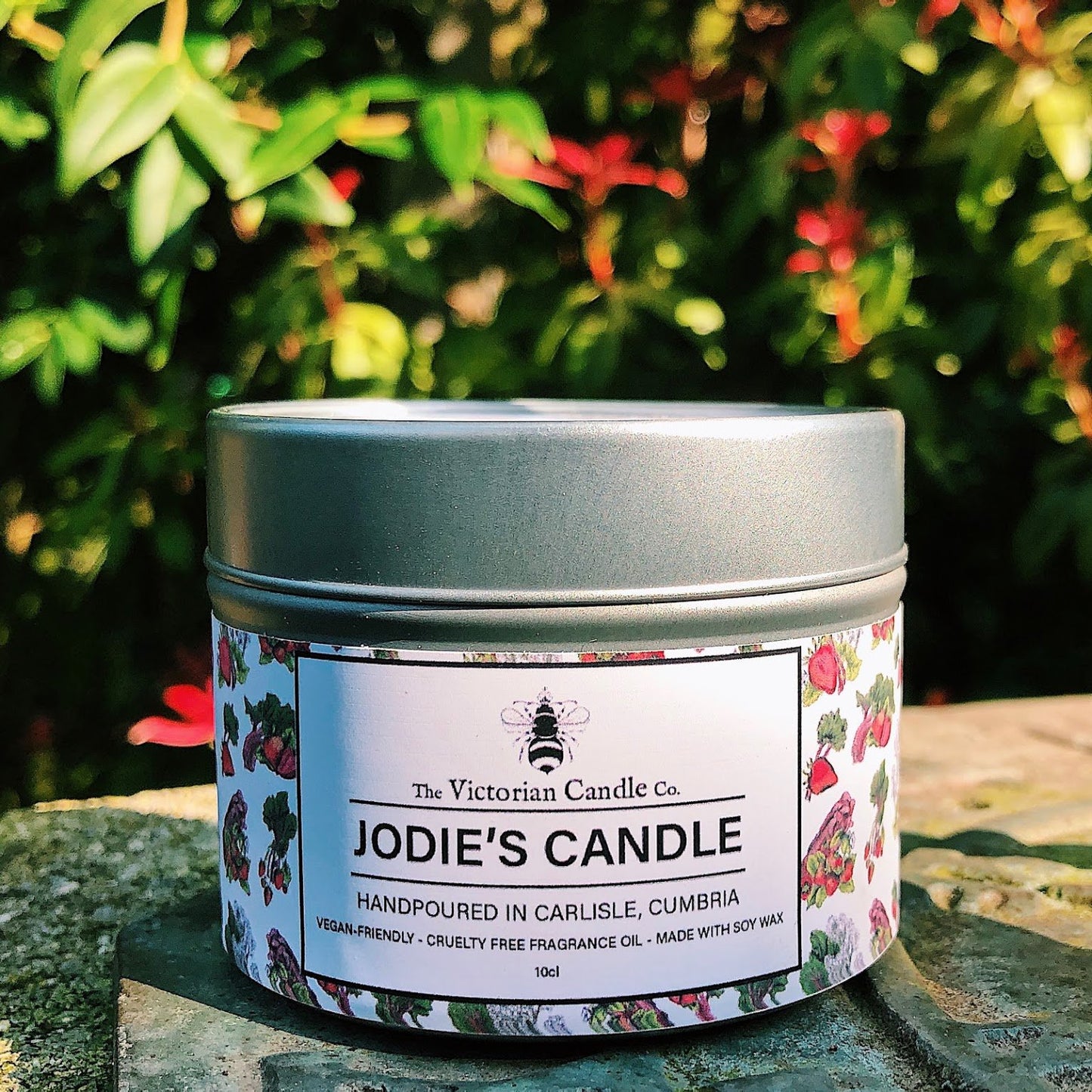 A candle tin labeled "Personalise your own candle!" from The Victorian Candle Co. rests outdoors on a stone surface, surrounded by lush green foliage and red flowers. Crafted from soy wax, this vegan-friendly and cruelty-free gem adds an elegant touch to any setting.