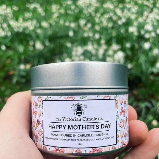 A hand holds a round 100ml tin candle from The Victorian Candle Co., labeled (Small) Black Cherry Mothers Day Limited Edition, featuring butterflies and flowers. In the background, a field of blurry white flowers sways gently.