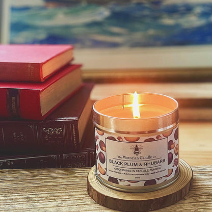 A lit (Large) Black Plum & Rhubarb Scented Candle in a 200ml tin from The Victorian Candle Co. rests on a wooden coaster, releasing its fruity aroma. Alongside it is a stack of red and brown hardcover books, and in the background, a painting with muted blues and greens enhances the serene ambiance.