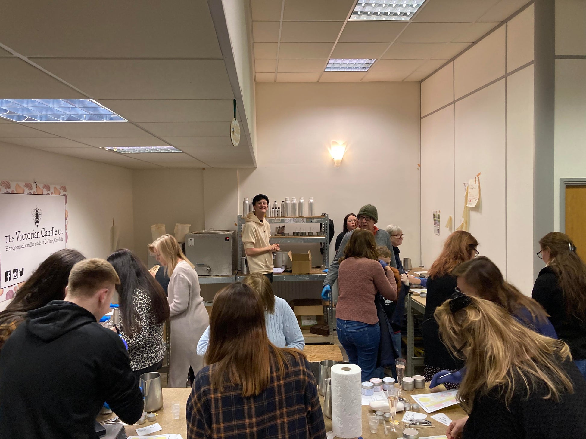 A group of people gathered in a Candle Making Workshops session, actively engaged in crafts, with supplies and materials spread across tables. The atmosphere is collaborative and focused, as a representative from The Victorian Candle Co. leads the session.