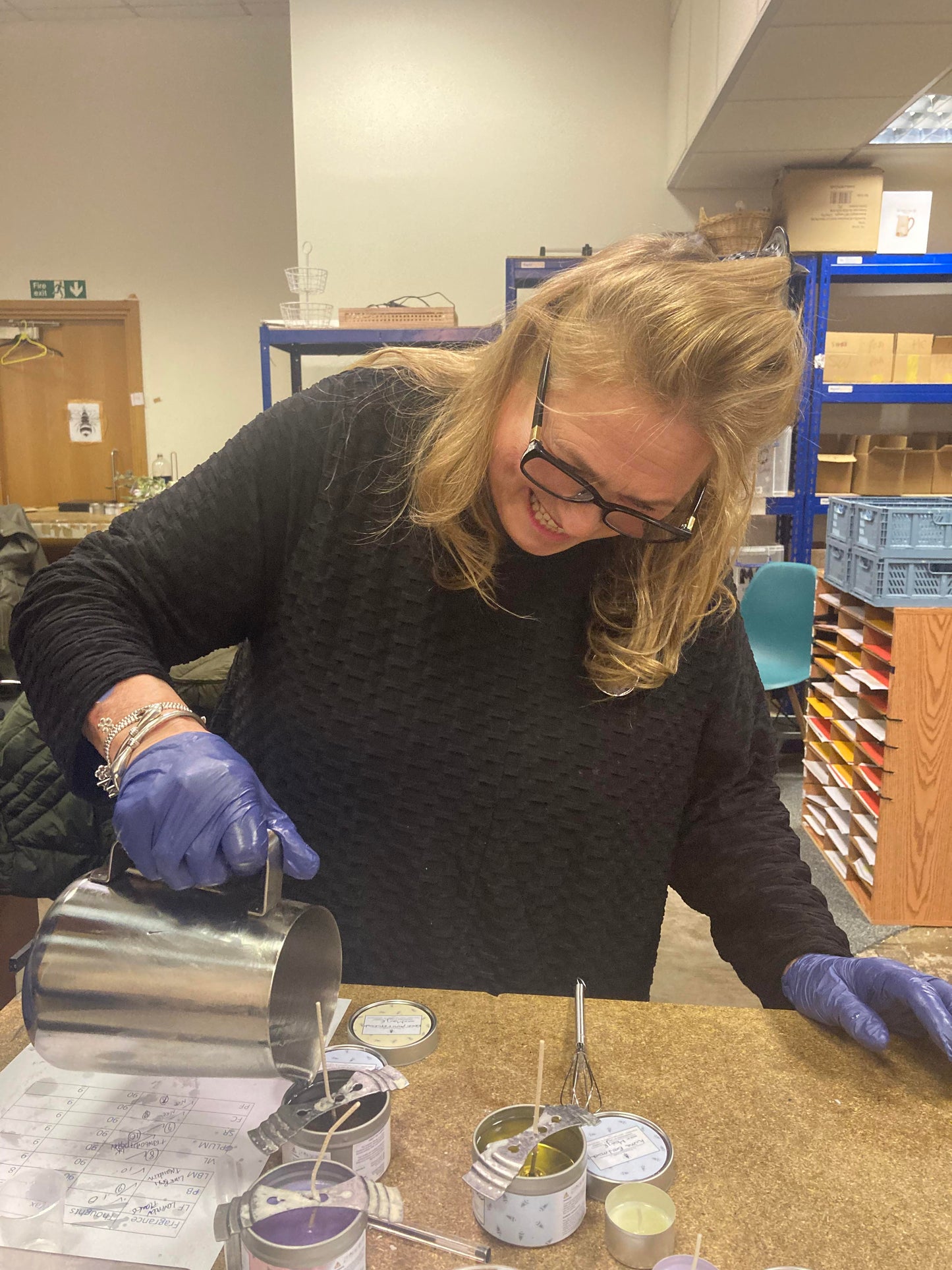 At The Victorian Candle Co., a woman with glasses and blue gloves, wearing a black sweater, smiles as she carefully pours wax from a metal jug into candle tins during a Candle Making Workshop. Shelves filled with supplies can be seen in the background, highlighting her immersion in the art of crafting candles.