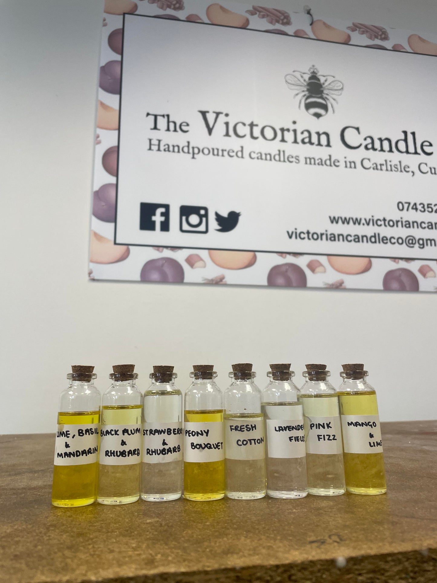 A row of small, labeled vials with various scented oils is showcased on a wooden surface, capturing the essence of a candle workshop. The labels highlight fragrances such as Lime, Basil & Mandarin and Lavender Fields. In the background, an elegant sign displays the name The Victorian Candle Co., further enhancing the scene.