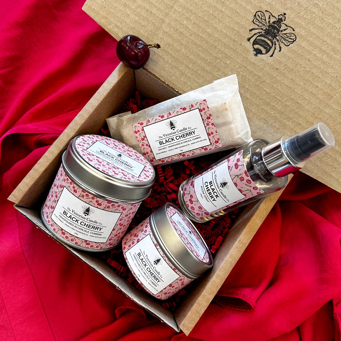 The Victorian Candle Co. Black Cherry Collection Box features a scented candle, body butter, soap, and room spray in pink cherry-patterned packaging with a cherry and bee design on the lid, displayed on red fabric.