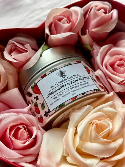 A heart-shaped box filled with light pink roses and a Strawberry & Pink Pepper scented candle from The Victorian Candle Co., elegantly presented in a 100ml metal tin. The label displays strawberries and states that it is hand-poured in Carlisle, Cumbria.