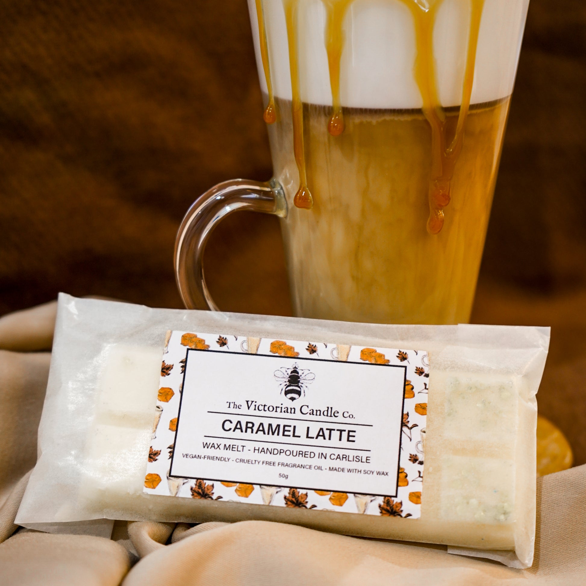 A glass mug of caramel latte sits behind The Victorian Candle Co. Caramel Latte - Wax Melt Snap Bar on beige fabric. The wax melt’s autumn leaf label evokes a cozy coffee scent, reminiscent of a fresh caramel latte.