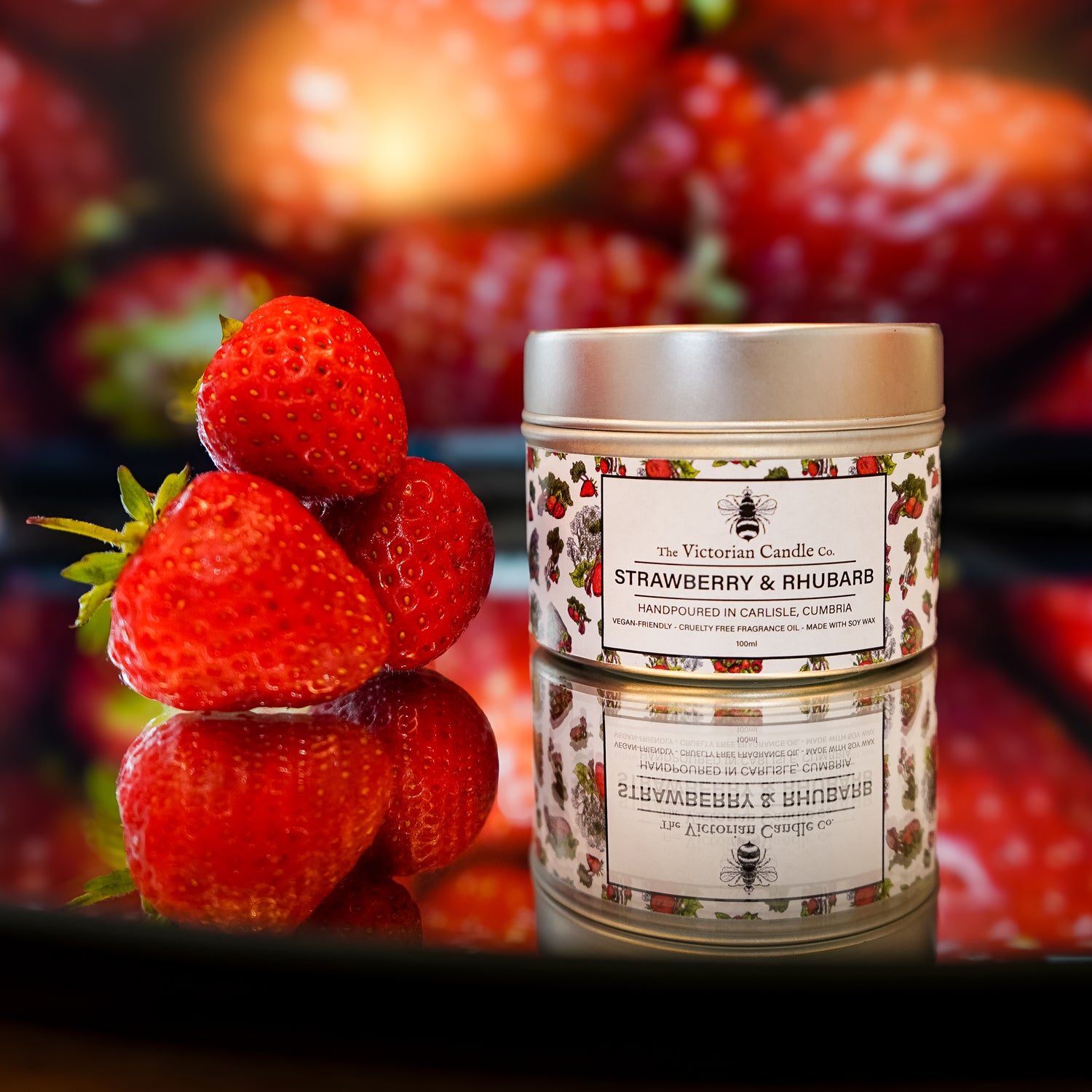A 100ml tin of The Victorian Candle Co. Strawberry & Rhubarb Scented Candle sits on a reflective surface beside three fresh strawberries, with more strawberries blurred in the background, showcasing this delightful soy wax candle.