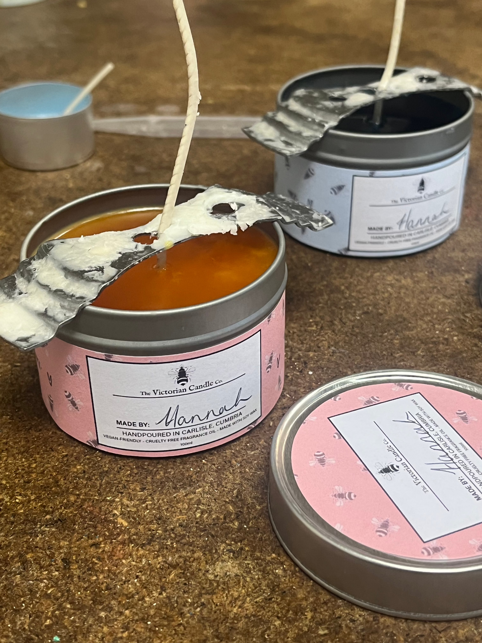 Three tin candles from The Victorian Candle Co.s Mothers Day Candle Making Workshops feature bestselling fragrances and rustic labels. Two display orange, layered designs with wicks, while one is topped with a silvery leaf, evoking a delightful workshop experience in Carlisle, Cumbria.