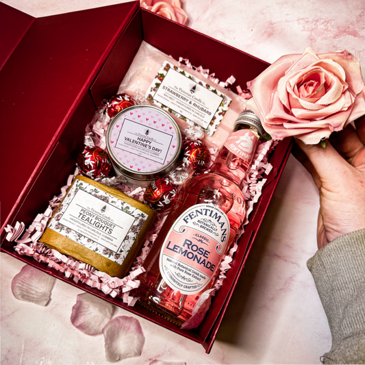 The Victorian Candle Co.s Valentine’s Day Deluxe Gift Box features pink wrapping, pink rose lemonade, bath salts, a Happy Valentines Day scented candle, Lindt Lindor Milk Chocolate Truffles, and a hand-held pink rose. Rose petals are gracefully scattered on the surface.