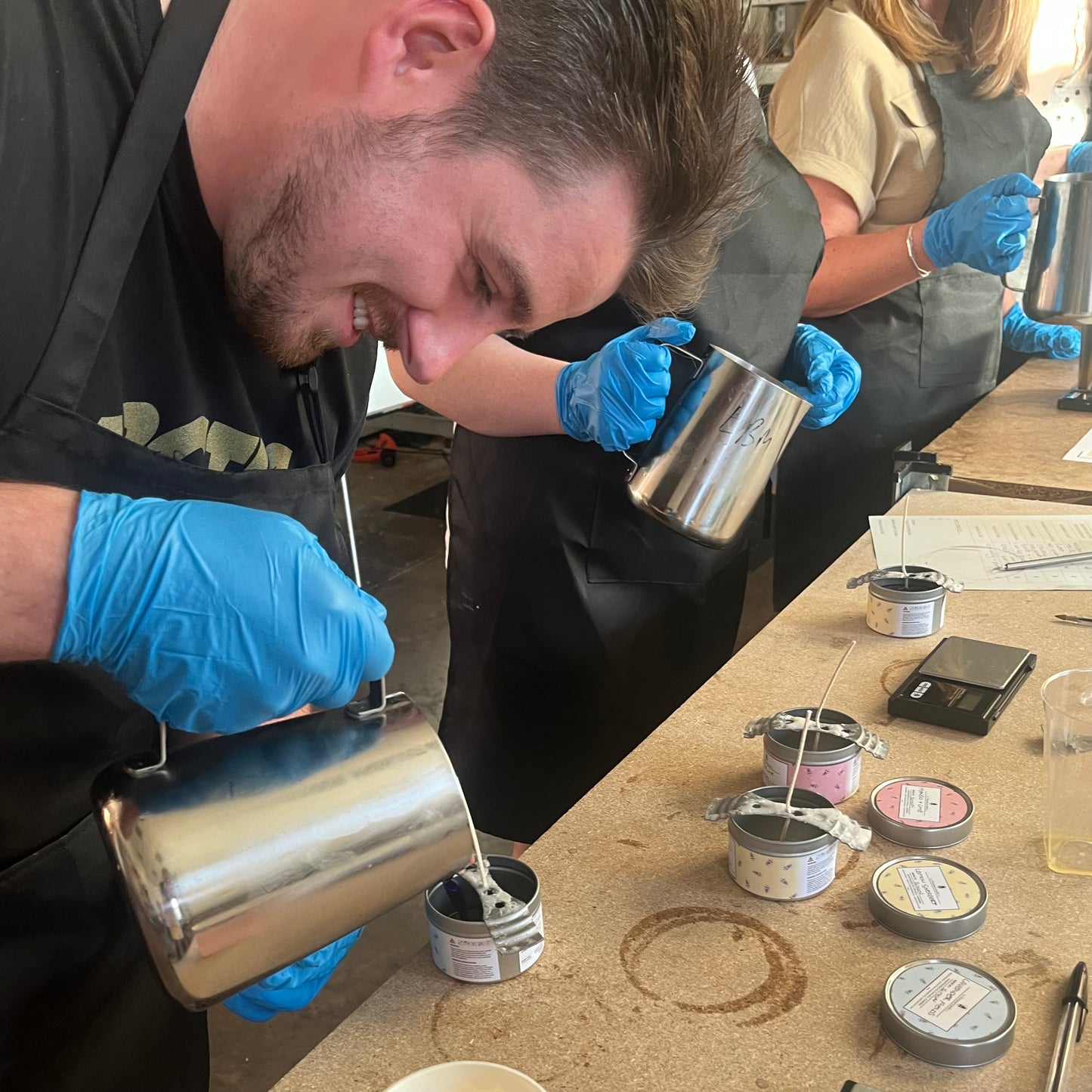 A man clad in blue gloves and a black apron carefully pours wax into a candle container during The Victorian Candle Co.'s Candle Making Workshops. Others nearby are fully absorbed in crafting candles, with various tins and tools scattered over the table, creating an atmosphere that is both focused and creative.