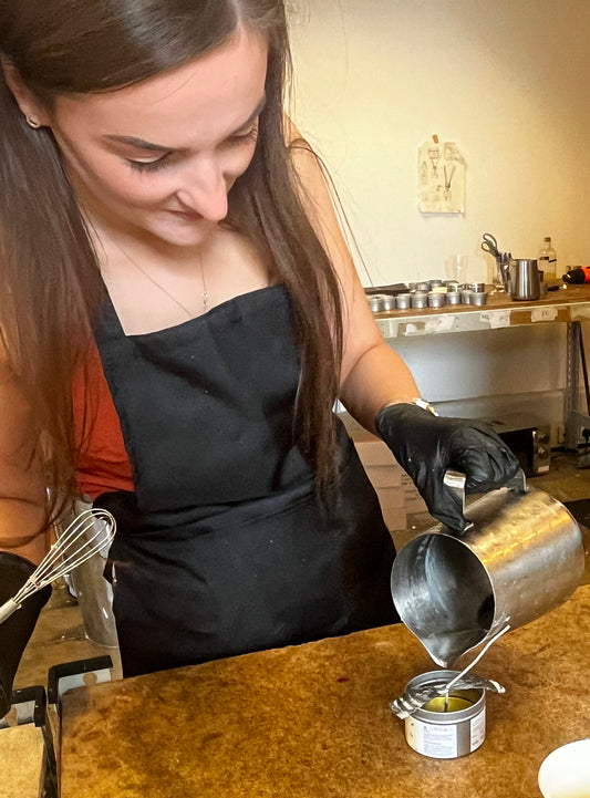 A woman in a black apron is focused on her task, carefully pouring liquid from a metal jug into a small container at The Victorian Candle Co.'s Candle Making Workshops. A whisk and various crafting materials surround her, showcasing the intricate process of candle making.